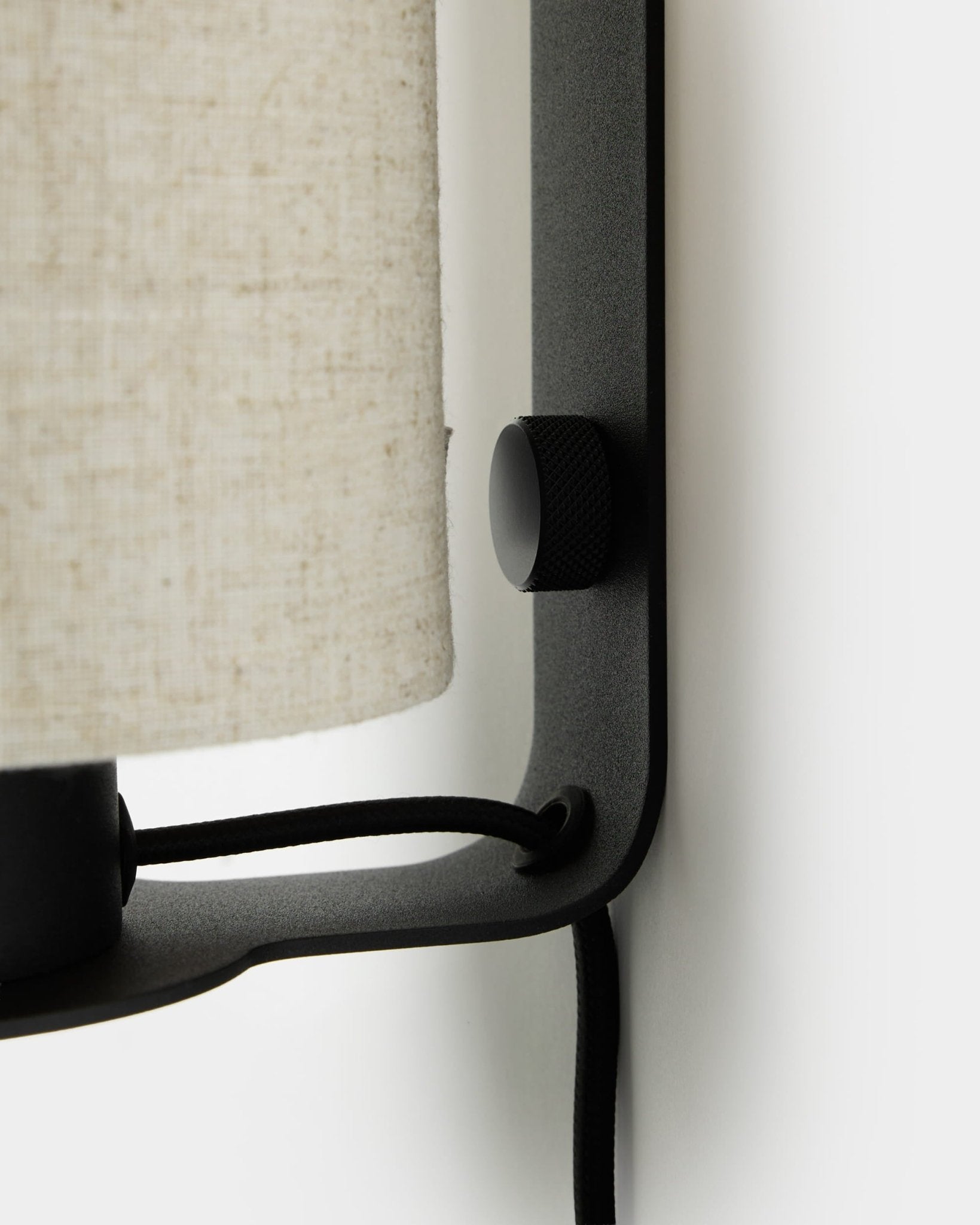 Närbild av WoodUpp Feya vägglampa i black och beige, med en texturerad ratt, skärm i linne-bomull och synlig kabel monterad på en vit vägg.