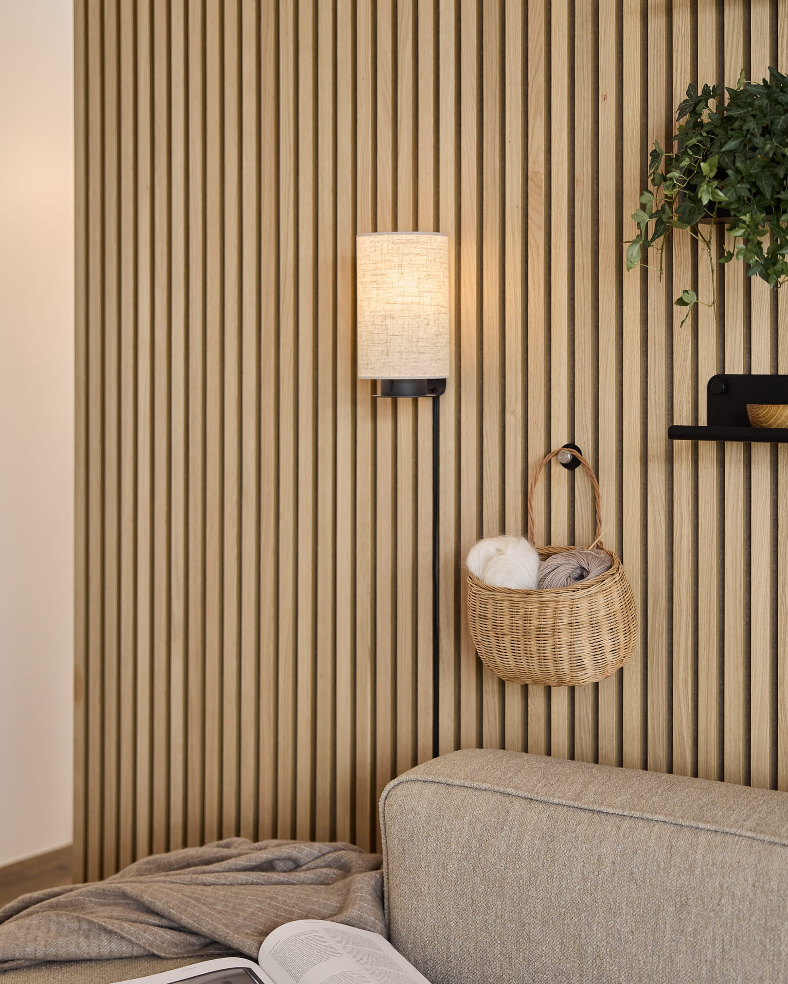 Beige couch next to a Nordic Akupanel wooden slat wall, with a woven yarn basket, wall lamp, plant on a shelf, and open book on gray blanket.