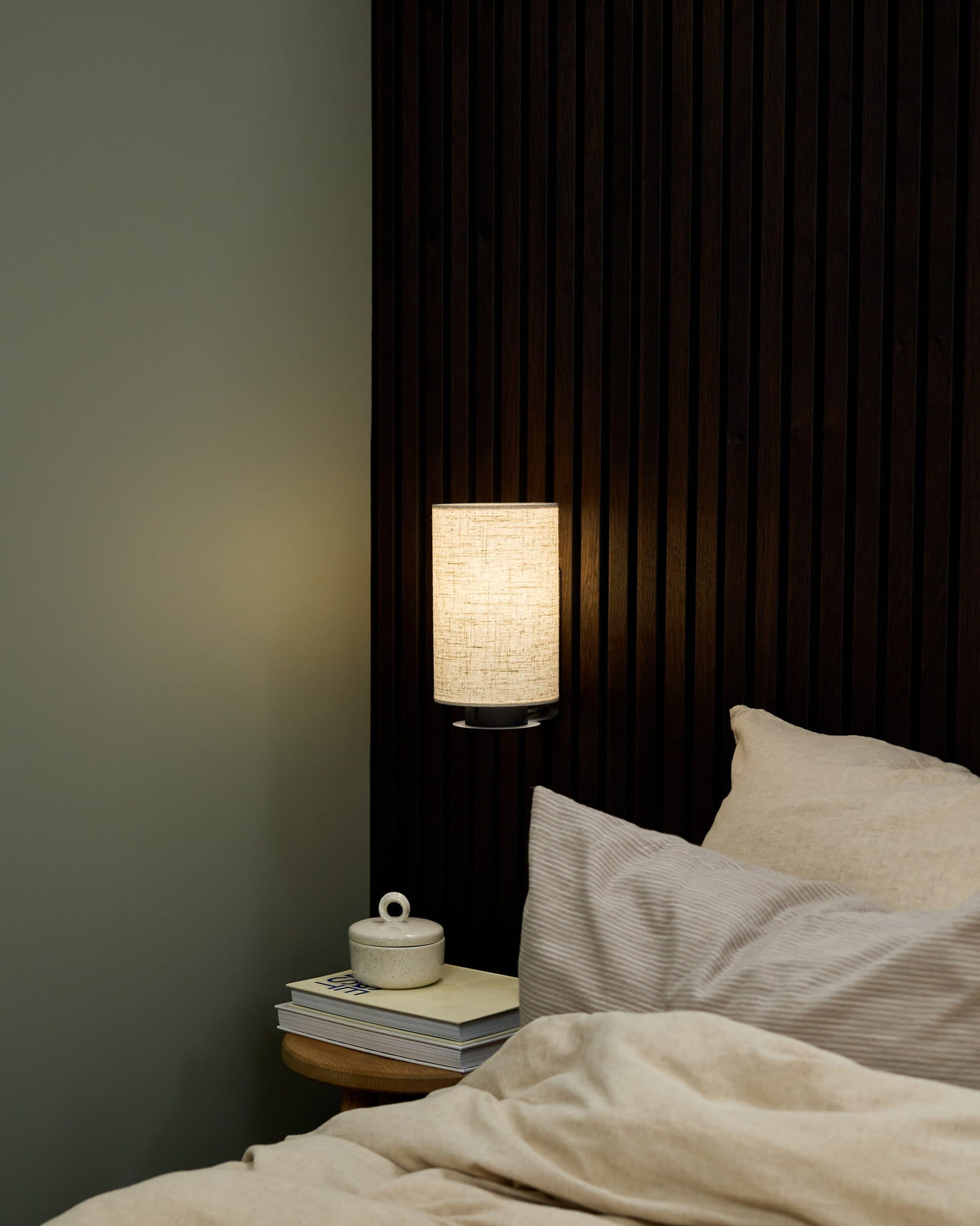 Sovrumshörna med WoodUpp Feya vägglampa i Black/Beige, sängkläder i beige och ett sängbord med böcker och en keramisk kopp med lock.