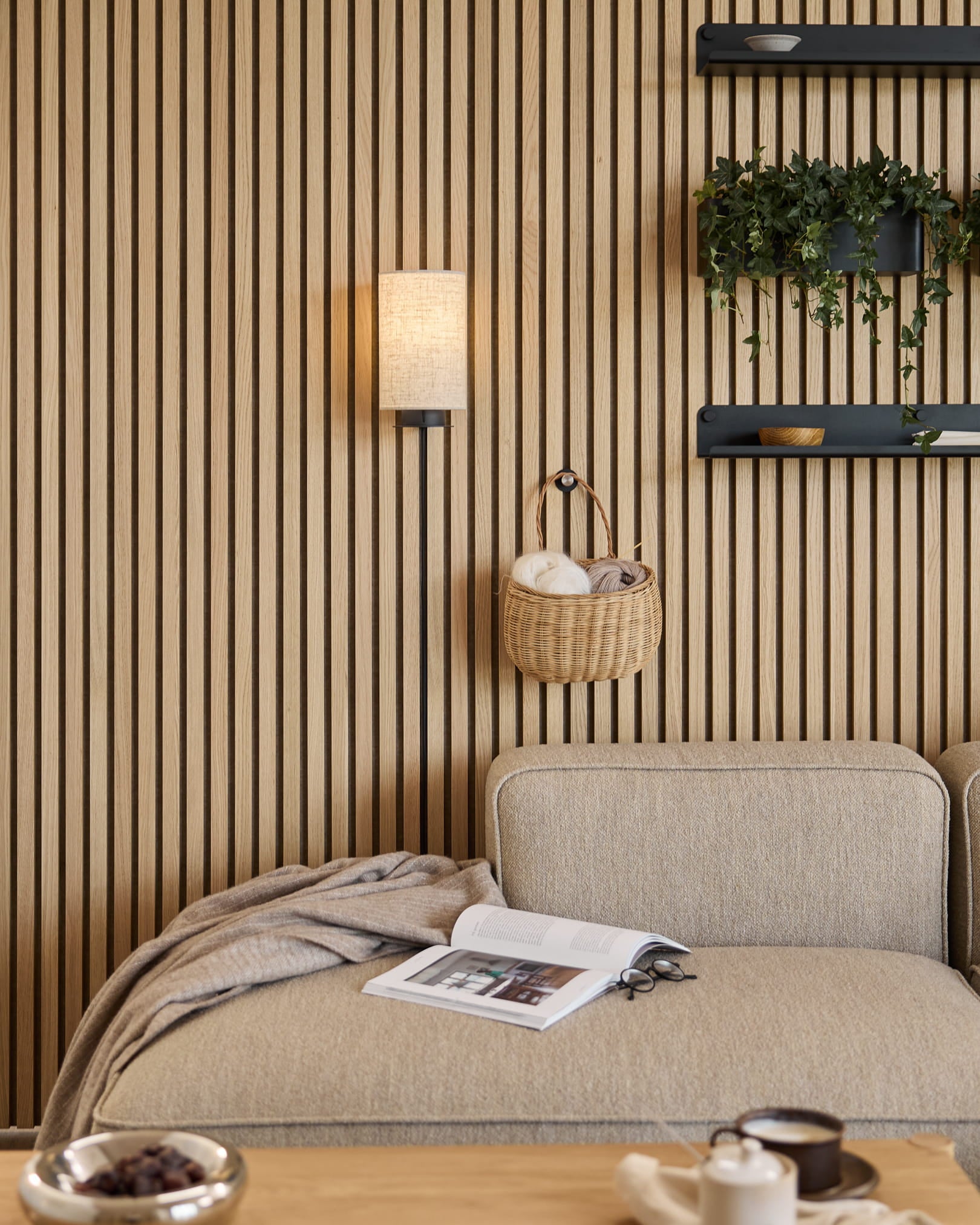 Beige soffa med pläd och bok intill en WoodUpp Akupanel akustisk ribbvägg, hyllor med växter och inredningsdetaljer samt en Feya vägglampa i Black/Beige.