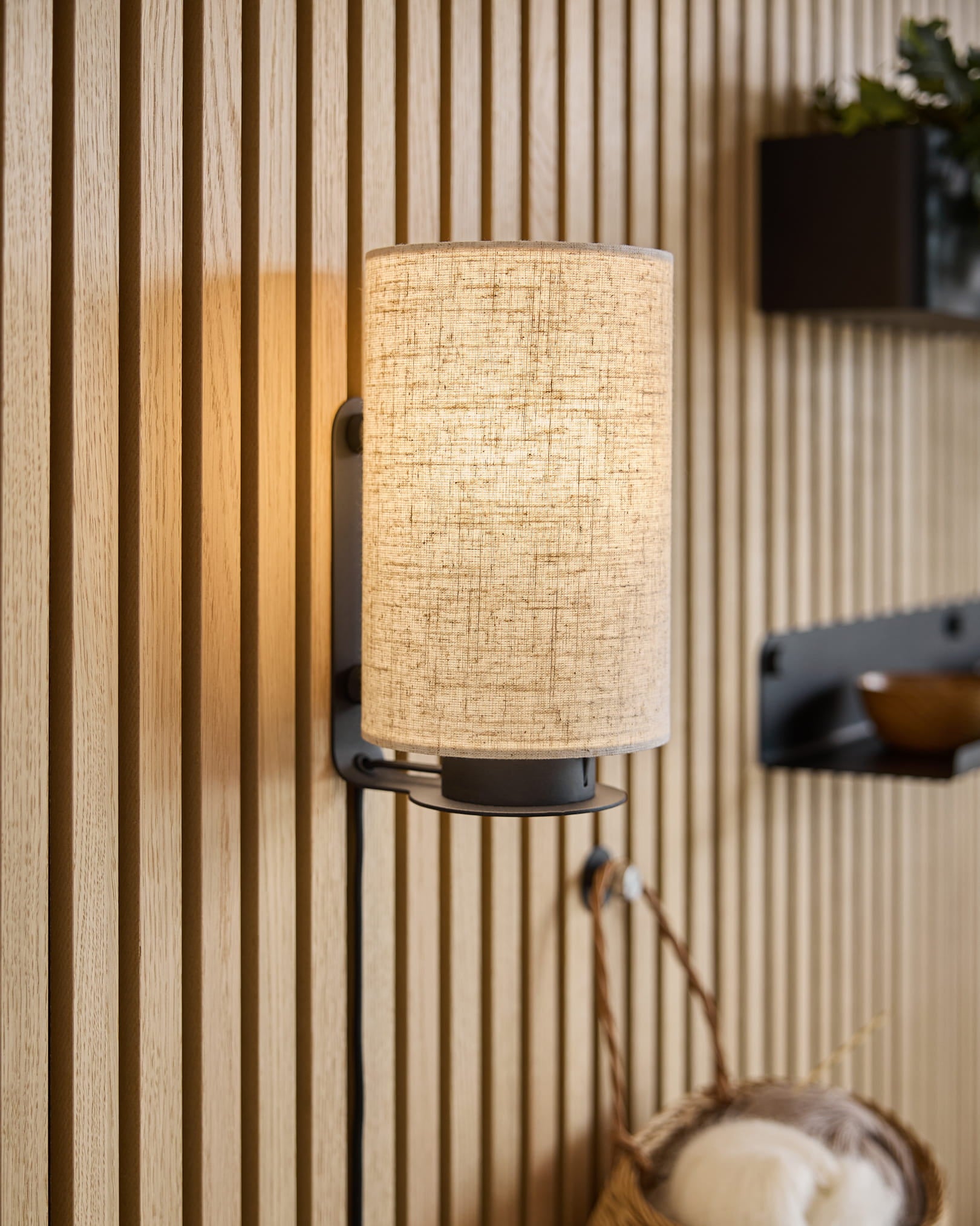 En modern vägglampa med tygskärm kastar varmt ljus på Akupanel akustiska träpaneler med textur, kompletterat med minimalistiska hyllor och en flätad korg.