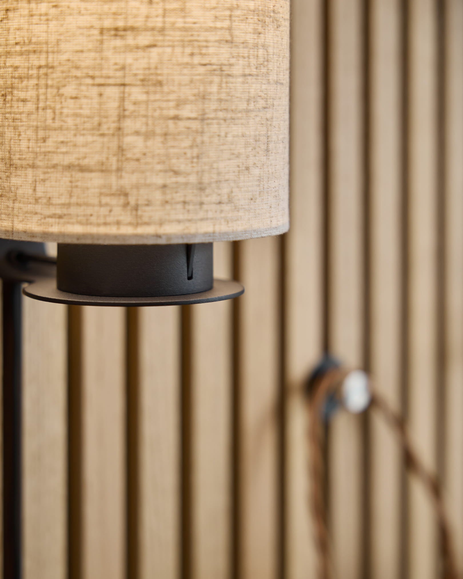 Feya vägglampa / Black/Beige från WoodUpp med en skärm i linne-bomull i beige, metallfot i black och synlig kabel monterad på en nordisk akustisk ribbväggspanel.