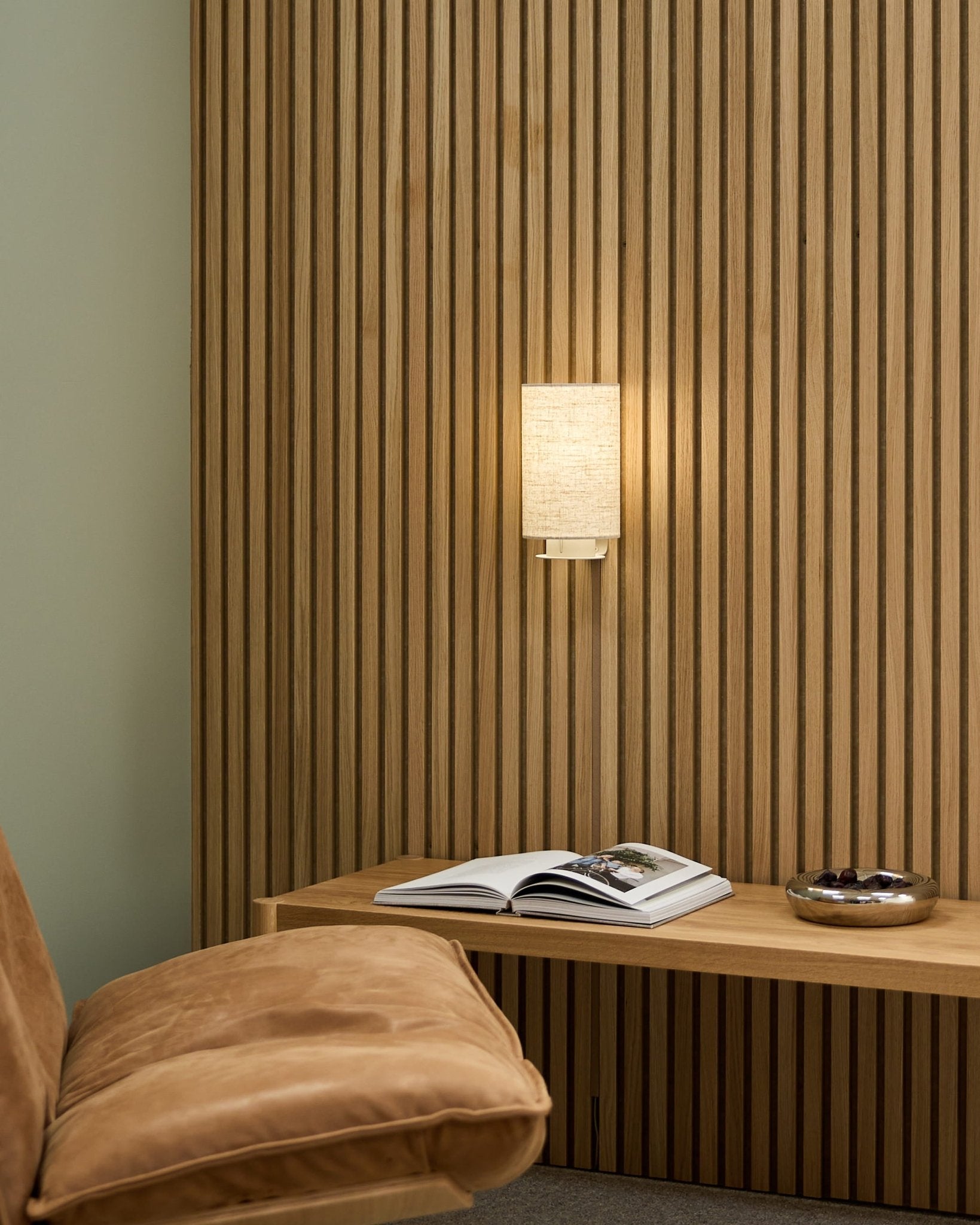 Minimalistiskt nordiskt rum med en brunbeige stol, WoodUpp Akupanel akustisk ribbvägg, Feya vägglampa i Beige/Beige, en uppslagen bok på en bänk och en skål.