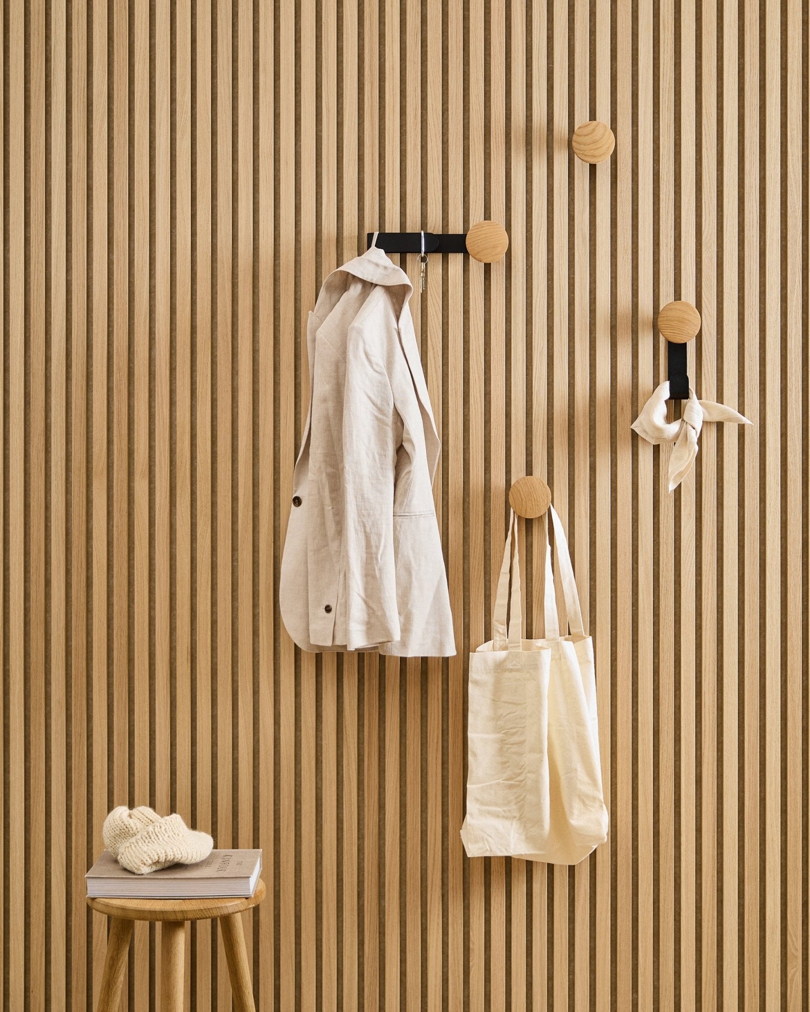 En beige jacka, tygväska och scarf hänger på WoodUpps Linn Horizontal Hanger i Natural Oak; en pall intill bär upp en bok och ett stickat plagg.