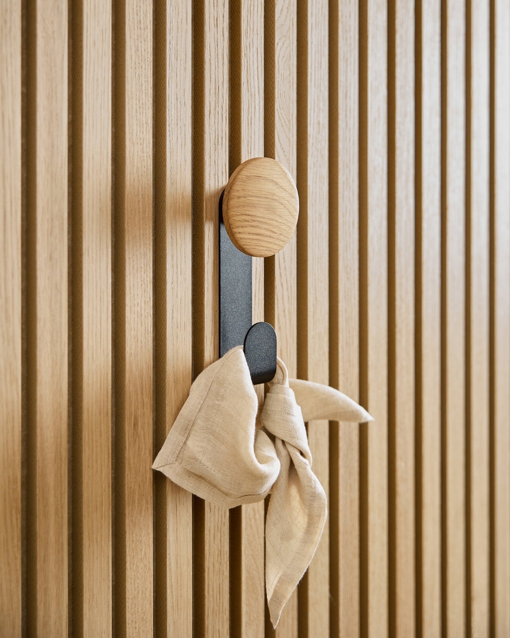 En beige handduk hänger på Linn Vertical Hanger i Natural Oak från WoodUpp, monterad på en vertical Akupanel akustisk ribbpanel.