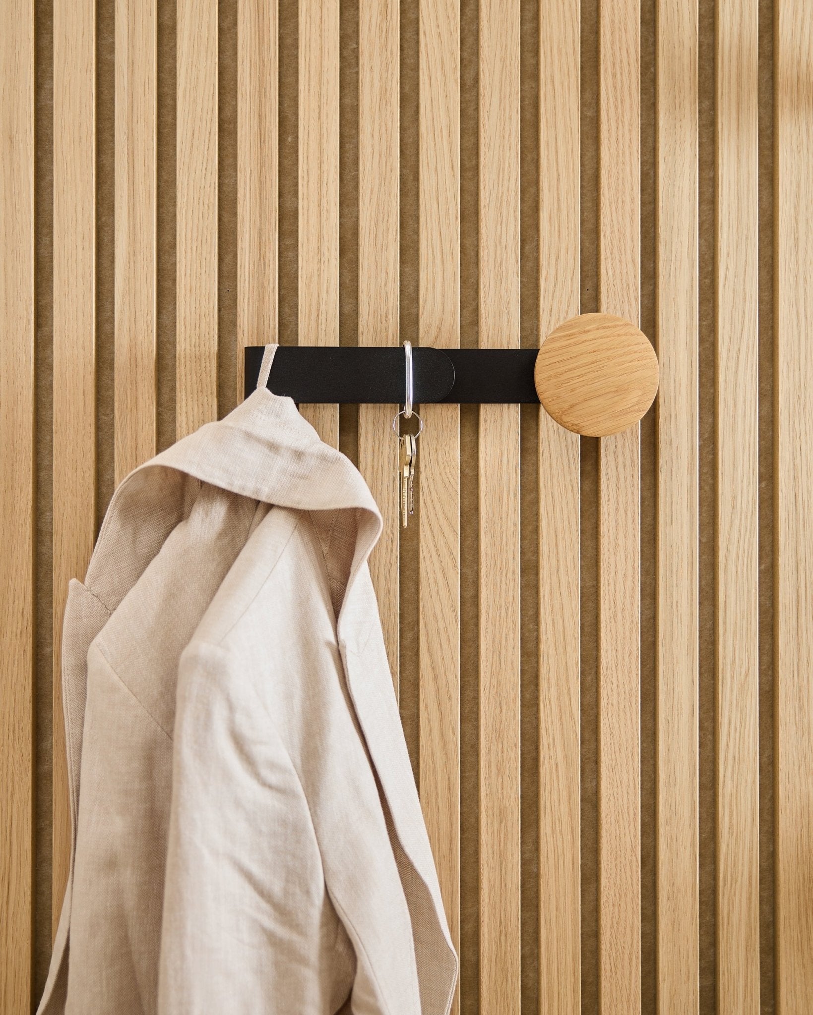 En beige kavaj och en uppsättning nycklar hänger på WoodUpp Linn Horizontal Hanger i natural oak, monterad på en natural oak Akupanel akustikpanel i trä.