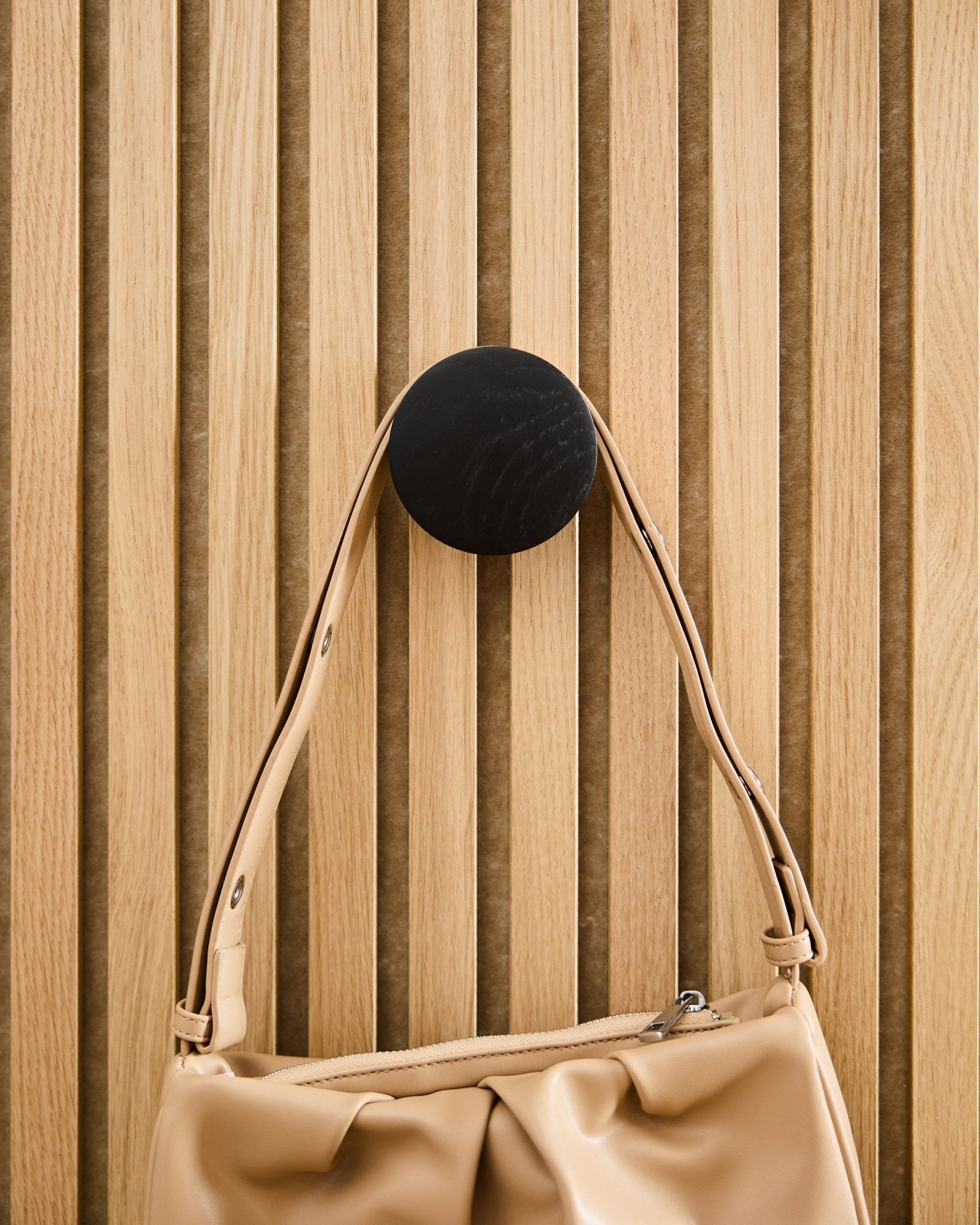 En Beige handväska hänger på en WoodUpp Linn | Single Hanger i Black Oak, monterad på en akustikpanel med ribbor i ljust trä Akupanel.