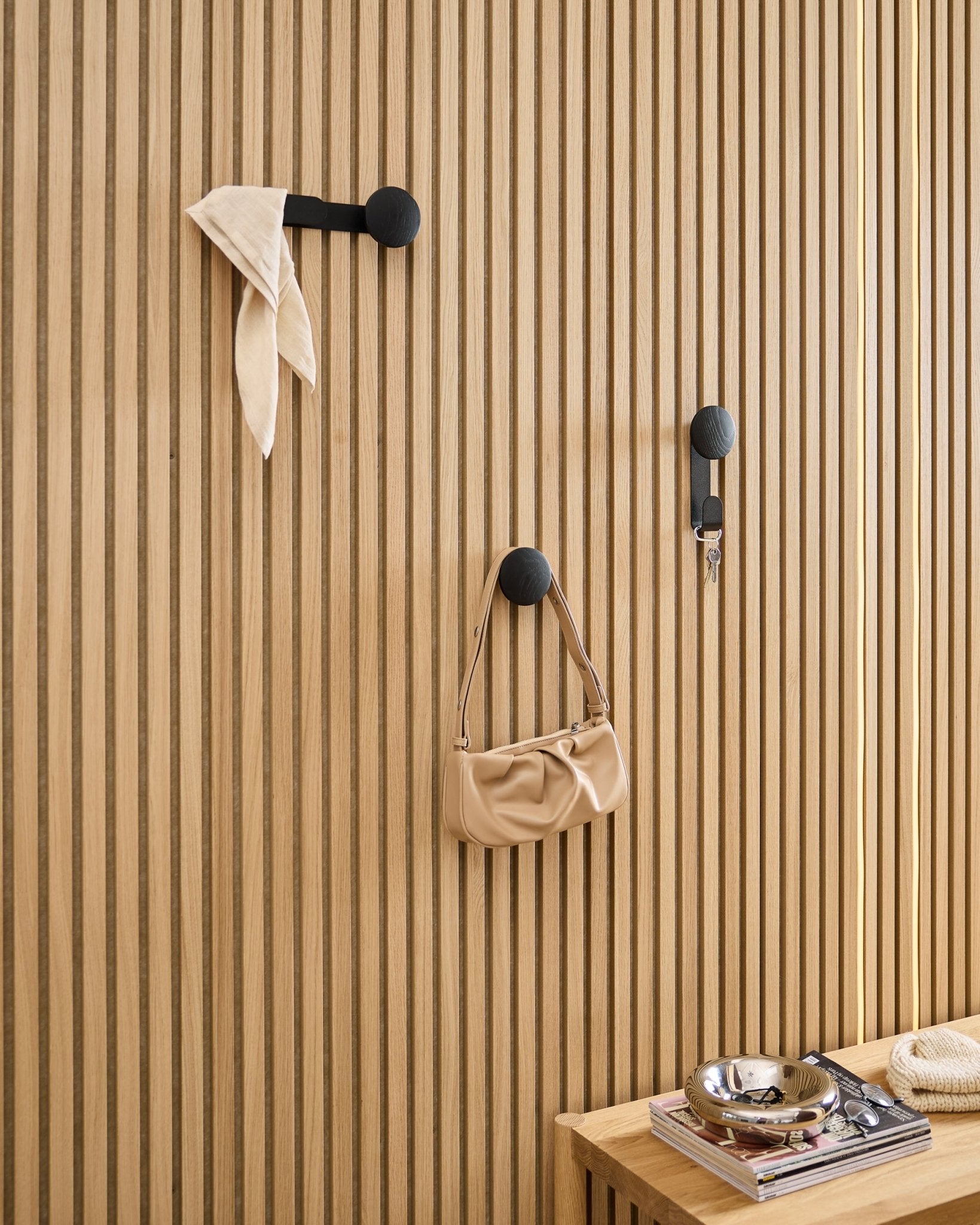WoodUpp Linn Single Hanger i Black Oak håller en scarf, beige väska och nycklar på en black Akupanel akustisk träpanel; bänk med böcker och metallfat i närheten.
