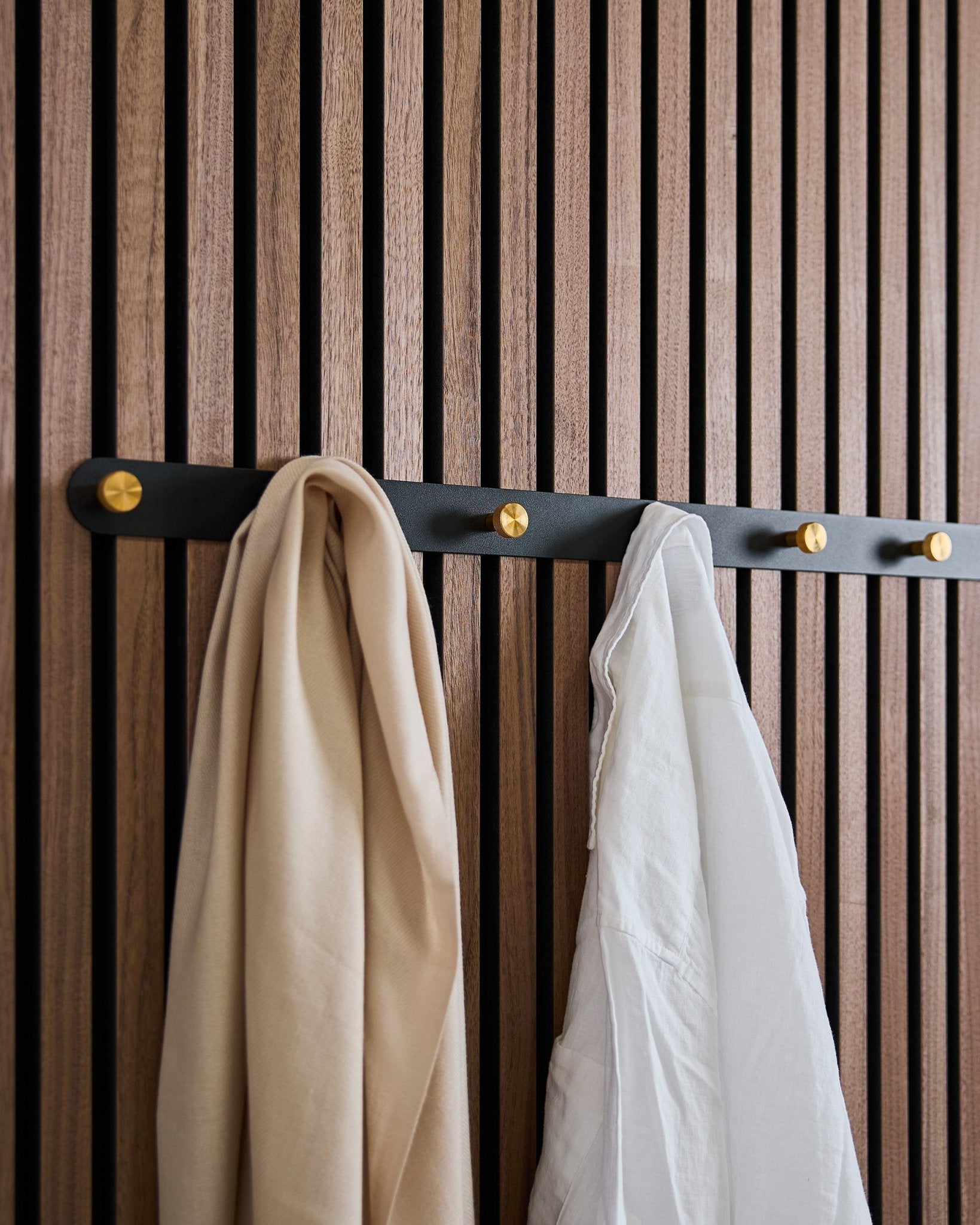 WoodUpp Norr Coat Hook Rack i black/brass (X7) med en beige scarf och vit skjorta hänger på en nordisk akustikpanel i trä med ribbor.