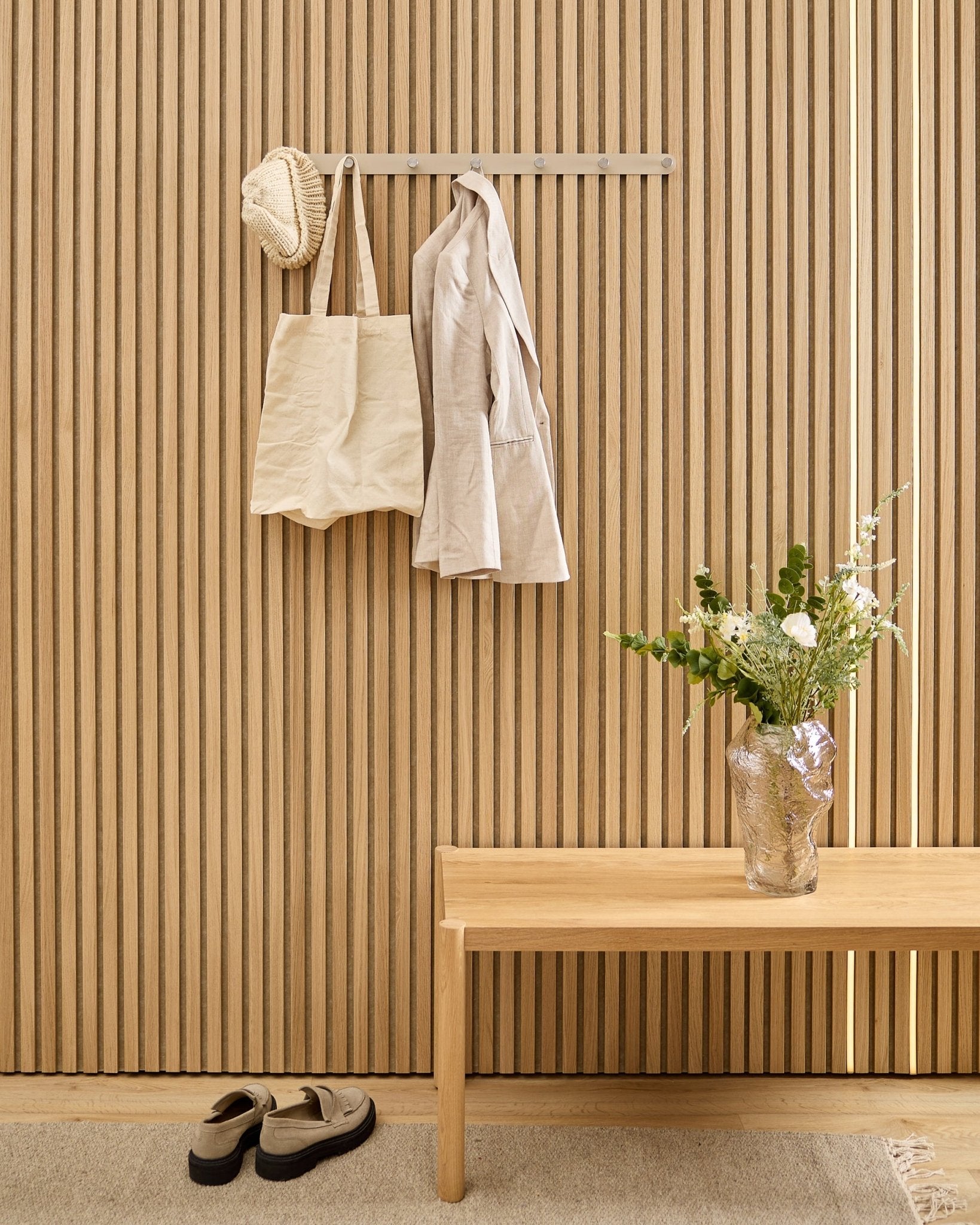 En beige väska, mössa och skjorta hänger på WoodUpp Norr klädhängare ovanför skor, med en bänk och vas bredvid den randiga akustikpanelen med trälameller.