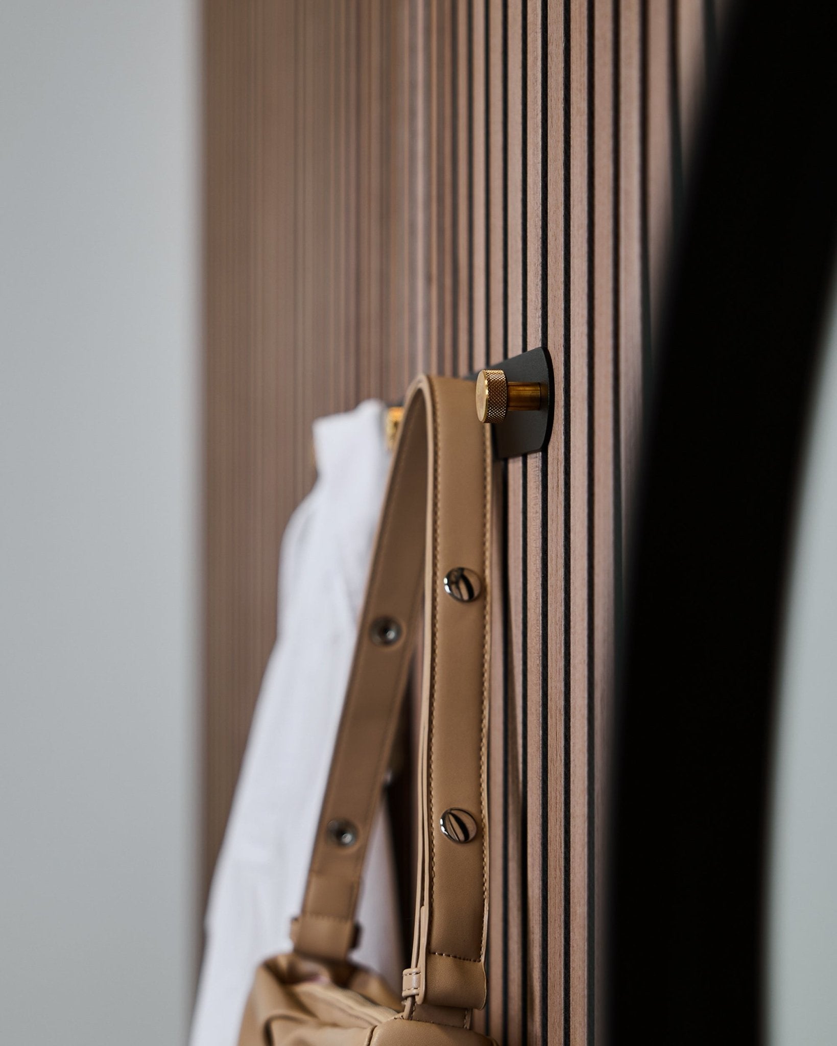 En ljusbrun väska och en vit skjorta hänger på en WoodUpp Norr knopplist (Black/Brass, X5) monterad på en vertical Akupanel akustisk ribbväggspanel.