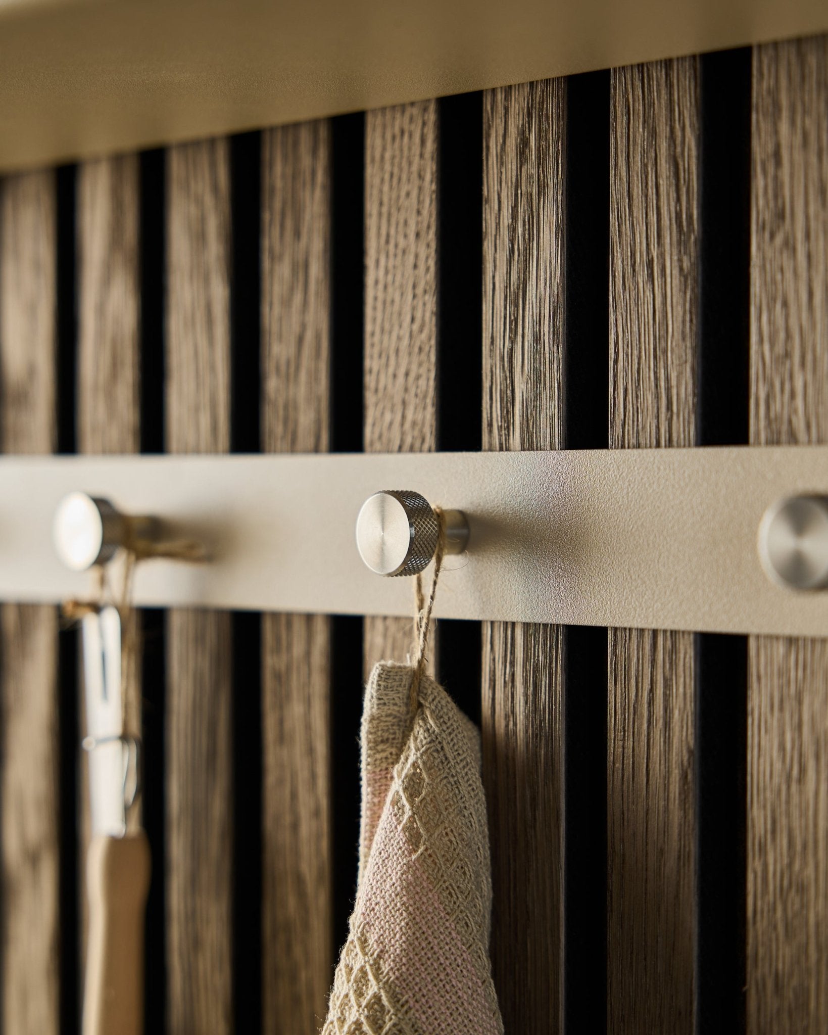 Närbild av WoodUpps Norr klädhängare (Beige/rostfritt Steel, X5) monterad på en ribbvägg i trä Akustikpanel, med en handduk och ett verktyg med handtag hängande på krokarna.