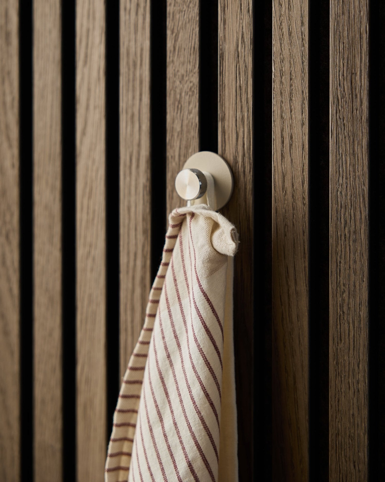 En randig handduk hänger på en beige Norr Coat Hook från WoodUpp, tillverkad i rostfritt steel, monterad på en texturerad akustisk spaltväggspanel i trä Akupanel.