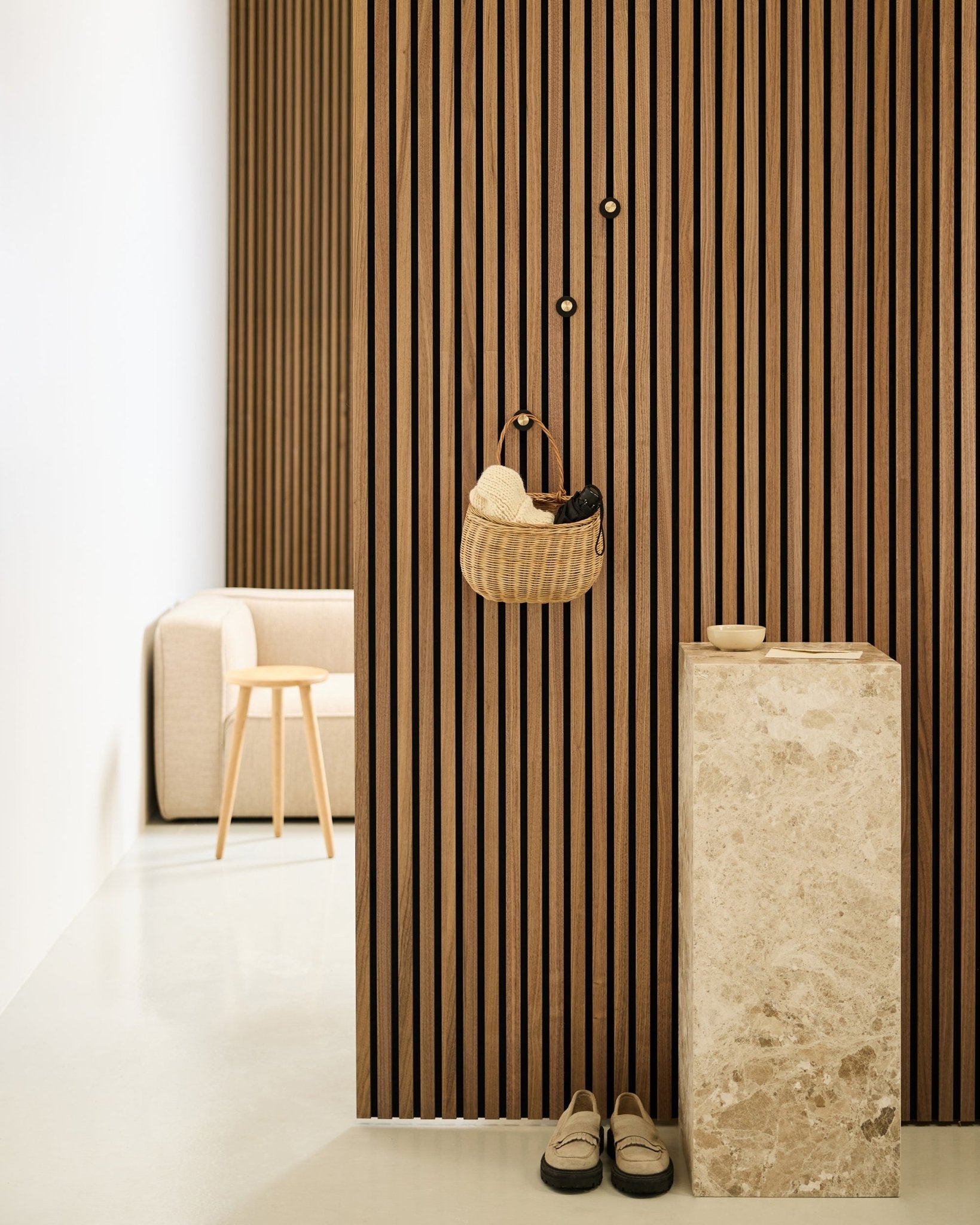Minimalistisk hall med Akupanel akustisk ribbvägg, WoodUpp Norr Coat Hook i black och brass, korg, stenpiedestal, tofflor och beige stol.