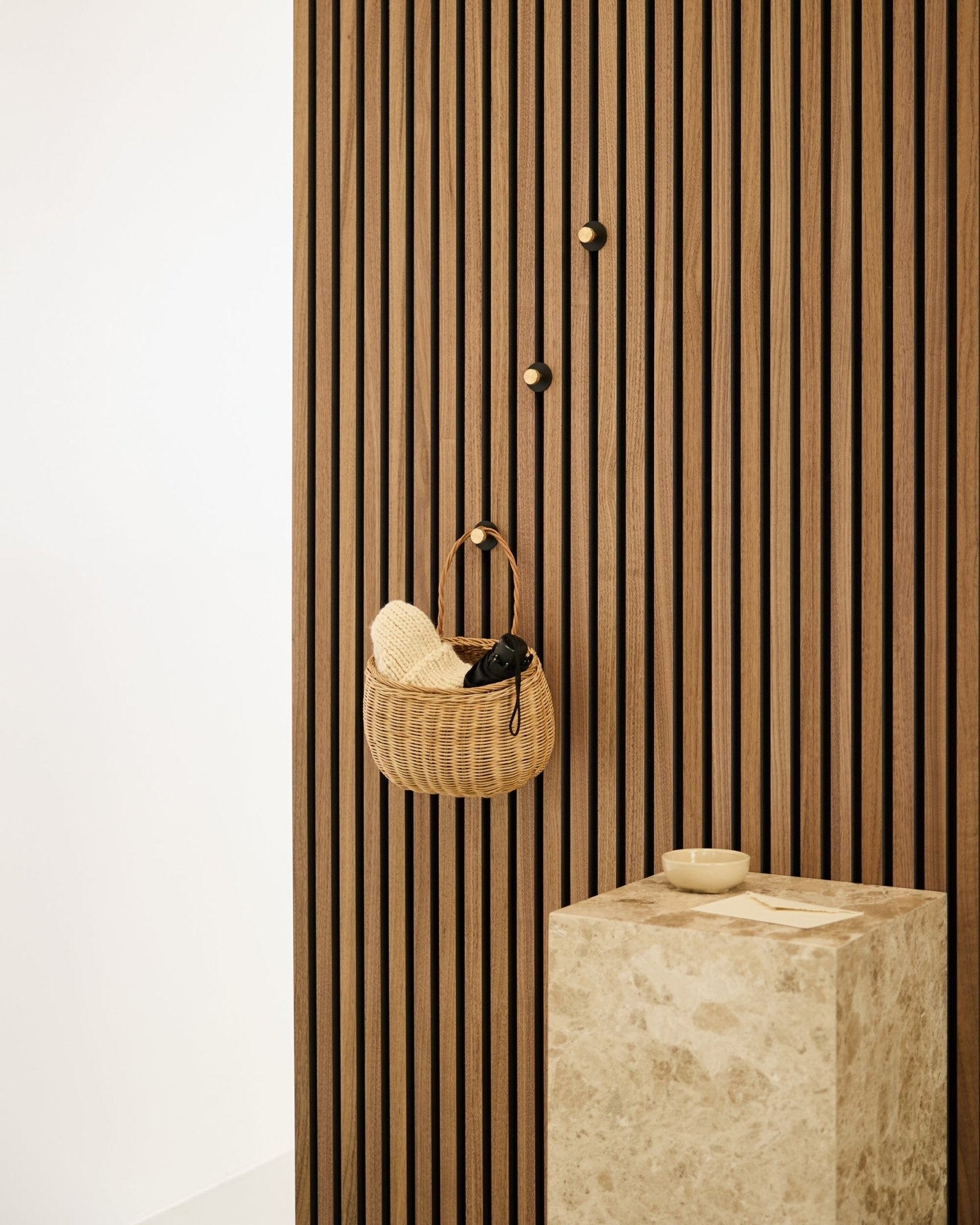 Norr Coat Hook från WoodUpp monterad på en nordisk Akupanel akustisk ribbvägg, bredvid en beige stenpiedestal med en small skål i en minimalistisk interiör.