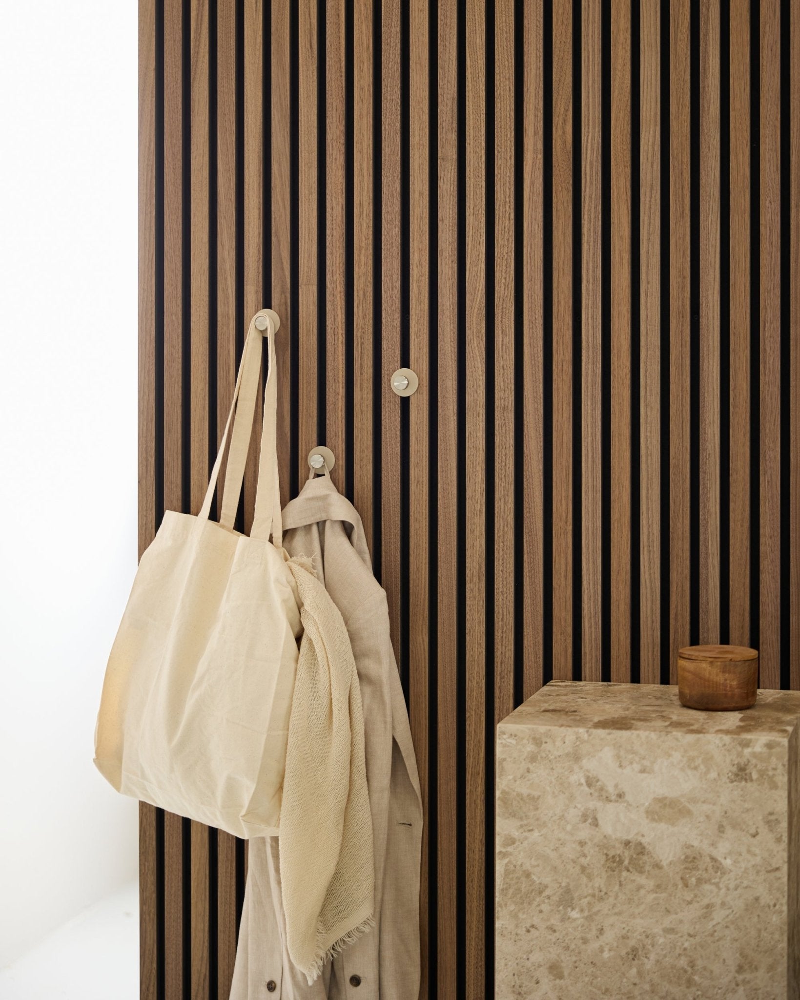 En beige tygväska och ett ljust tyg hänger på en WoodUpp Norr Coat Hook (Beige/rostfritt Steel) monterad på en vertical {{957f8f2c07fd8674387d91fad8e734b1f}} Akustikpanel bredvid en stenyta.