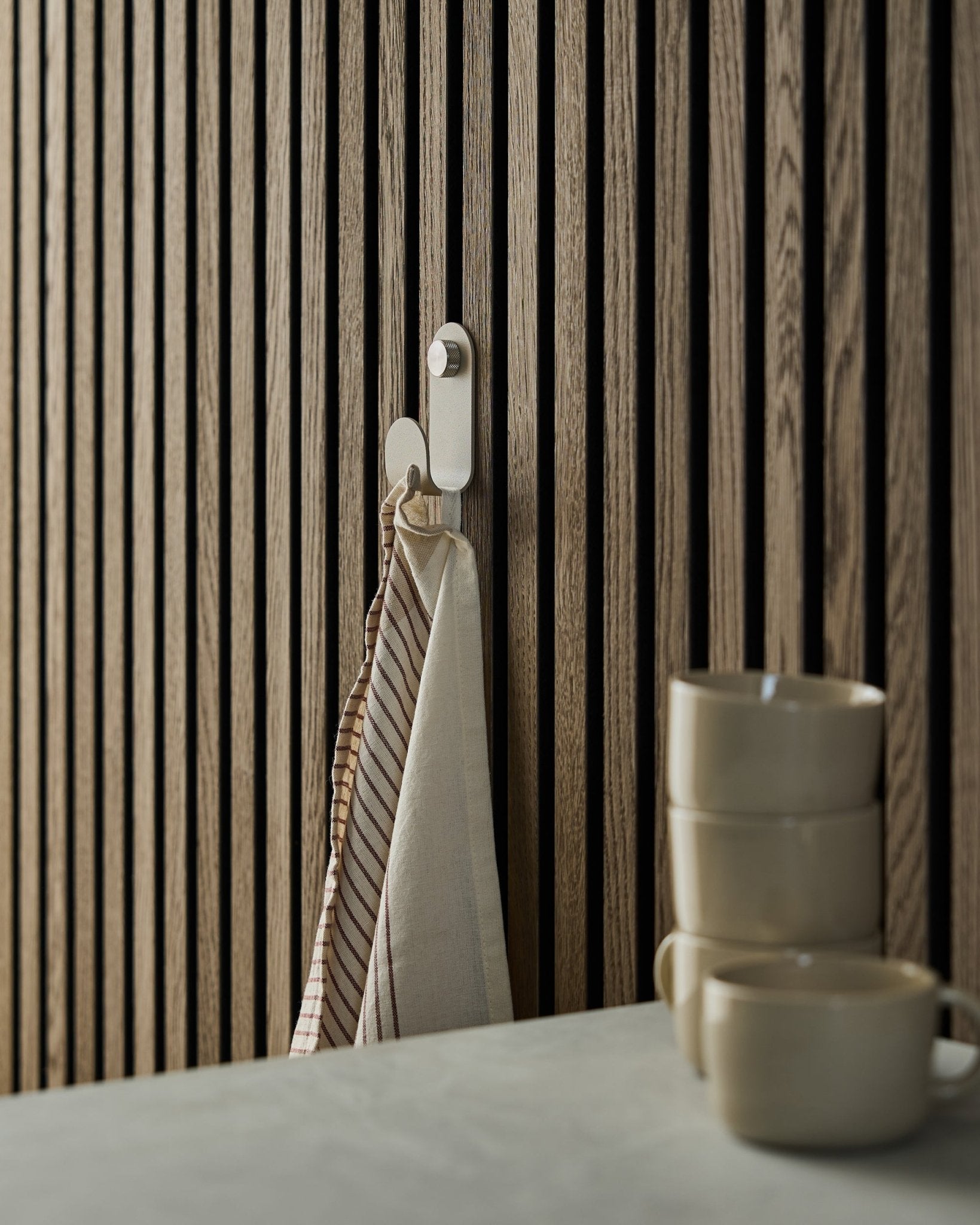 En randig handduk hänger på en WoodUpp Norr Single Hanger i beige, monterad på en akustisk ribbväggspanel i nordisk stil; tre beige muggar är staplade framför.