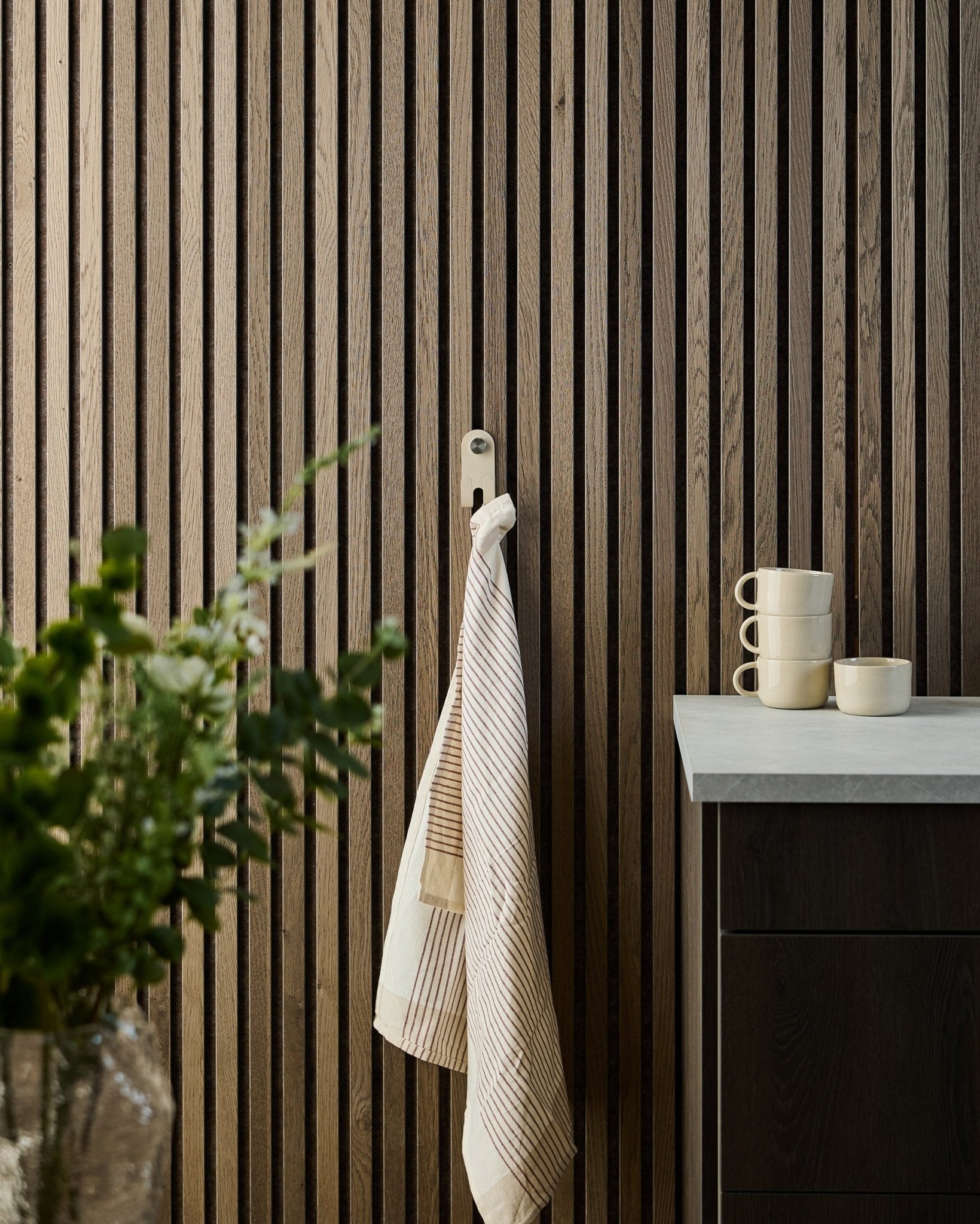 En randig handduk hänger på WoodUpp Norr Double Hanger i beige, monterad på en nordisk akustisk ribbväggspanel; muggar, en skål och en grön växt står nedanför.