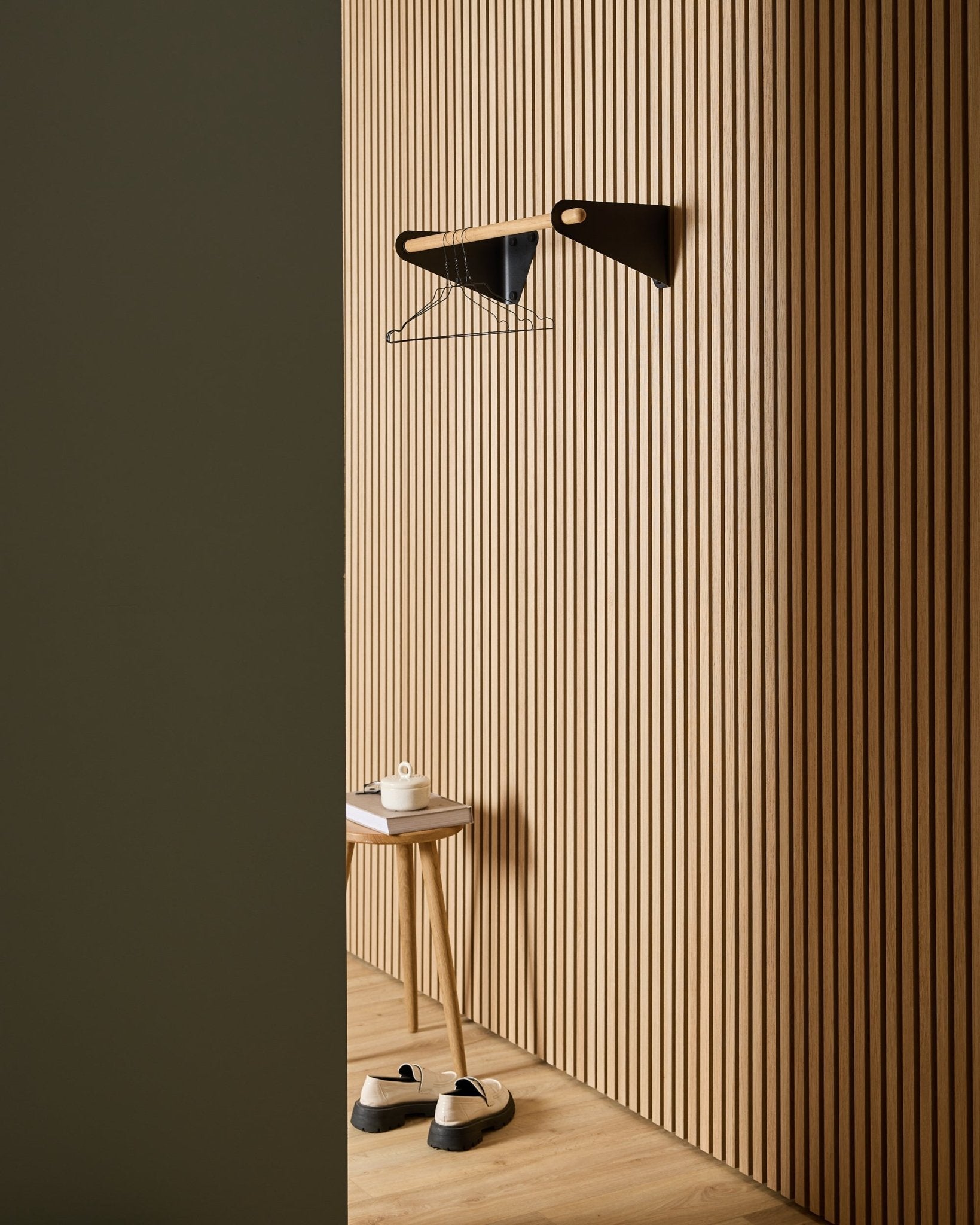 Minimalistisk hall med WoodUpp Akupanel nordiska akustikpaneler i träspjälor, black Linn Coat Rail (75,9 cm), pall, skor, vägghylla och pappersrulle.