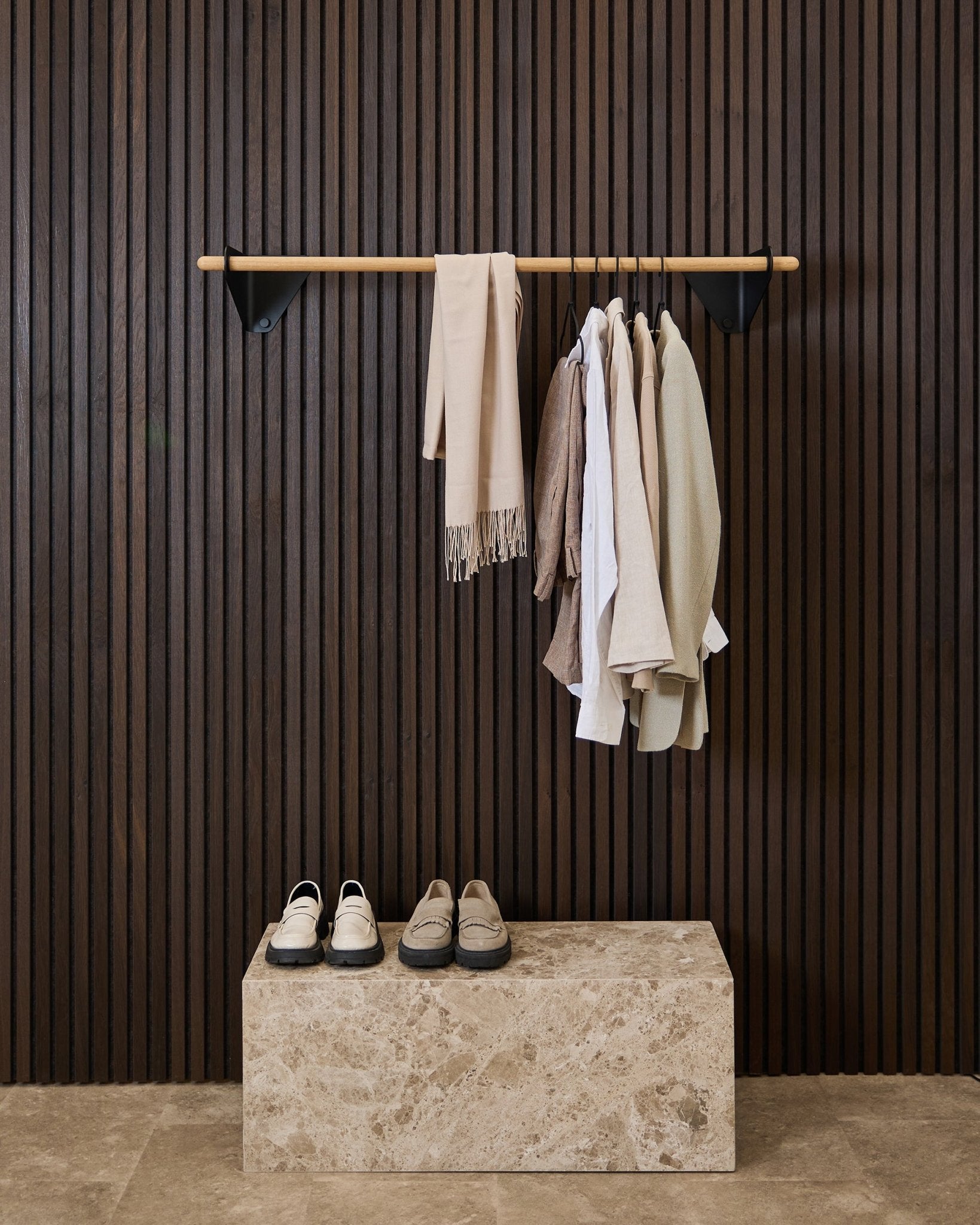 Kläder i neutrala toner och en scarf hänger på en black 115,9 cm Linn | Coat Rail från WoodUpp ovanför en bänk i marmor, mot en mörk Akupanel akustikpanel med ribbor.