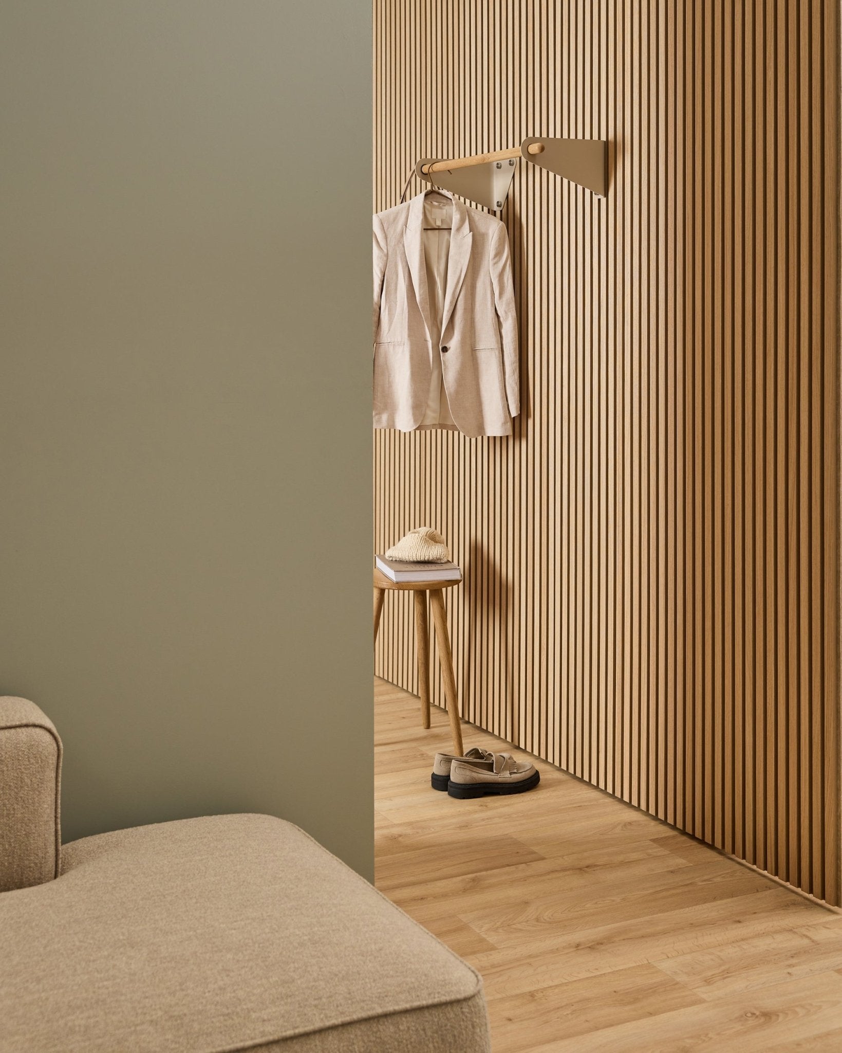 Minimalistiskt rum med beige soffa, Akupanel akustisk ribbvägg, WoodUpp Linn Coat Rail i beige (75,9 cm), pall med hatt och skor på ljust trägolv.