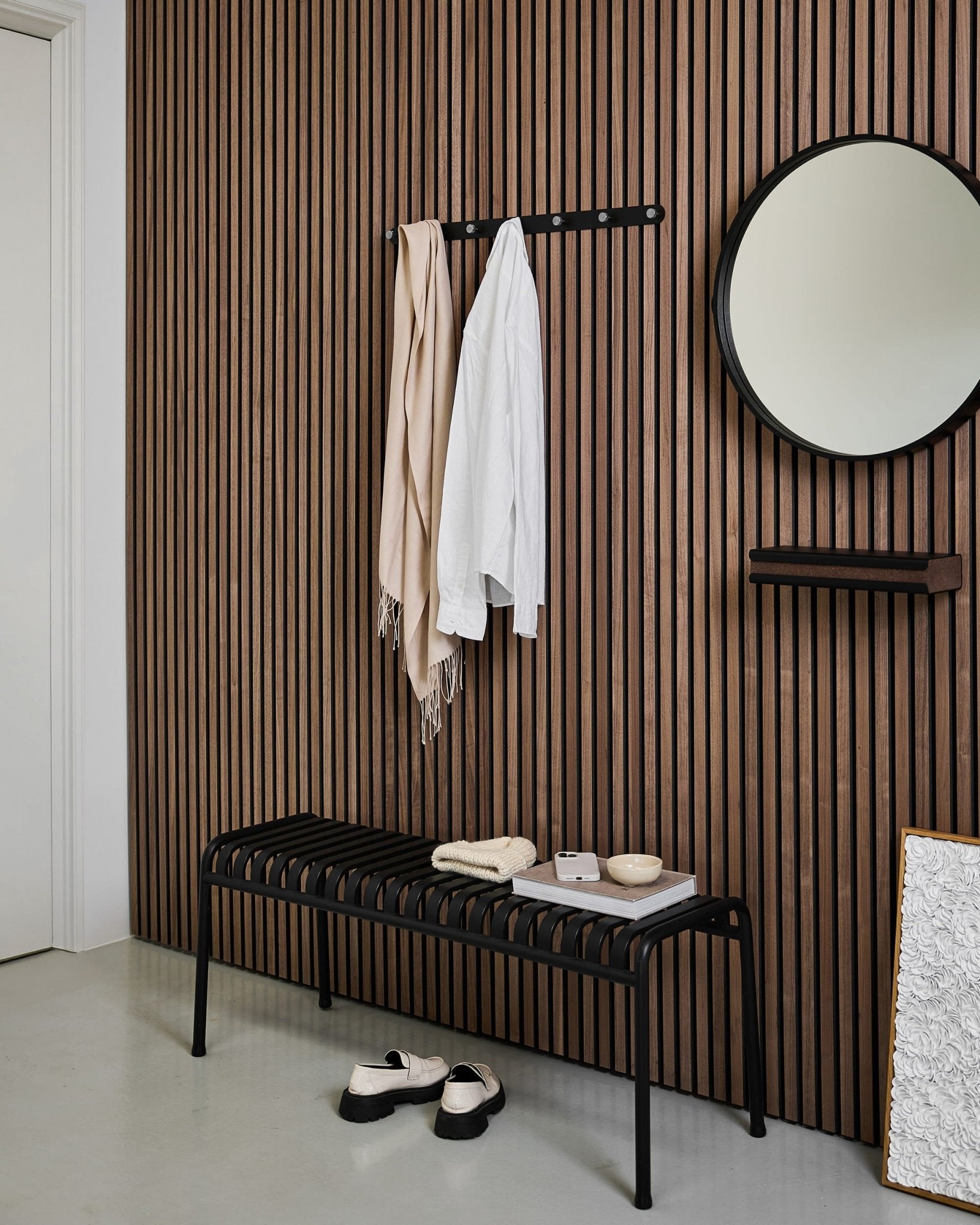 Modern entré med en nordisk akustikpanelvägg i ribbat trä, rund spegel, black bänk, WoodUpp Norr Coat Hook Rack och tofflor på ett ljust golv.