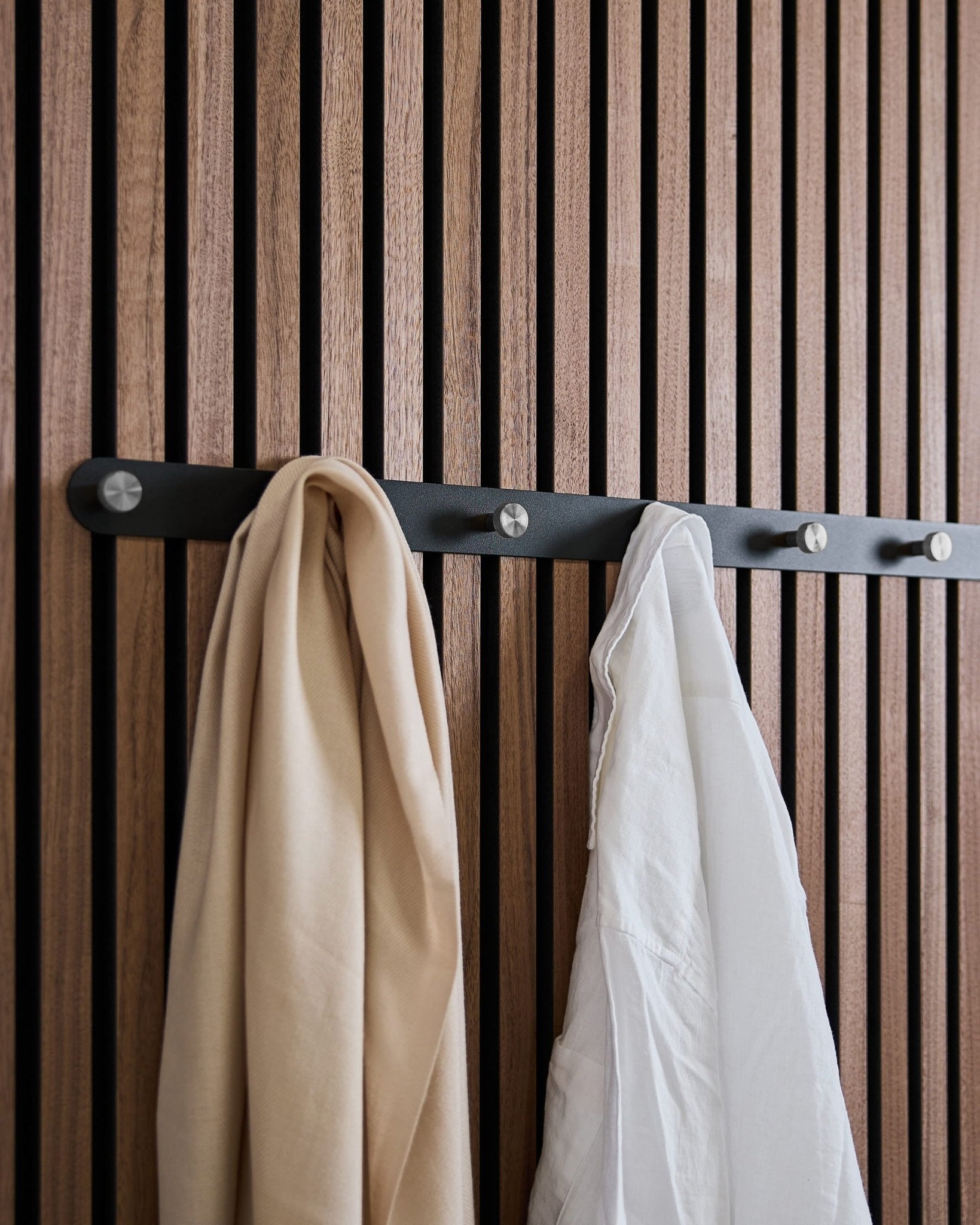 En beige scarf och vit skjorta hänger på WoodUpps Norr X7 Black/Stainless Steel klädhängare, monterad på en nordisk akustisk ribbväggspanel.