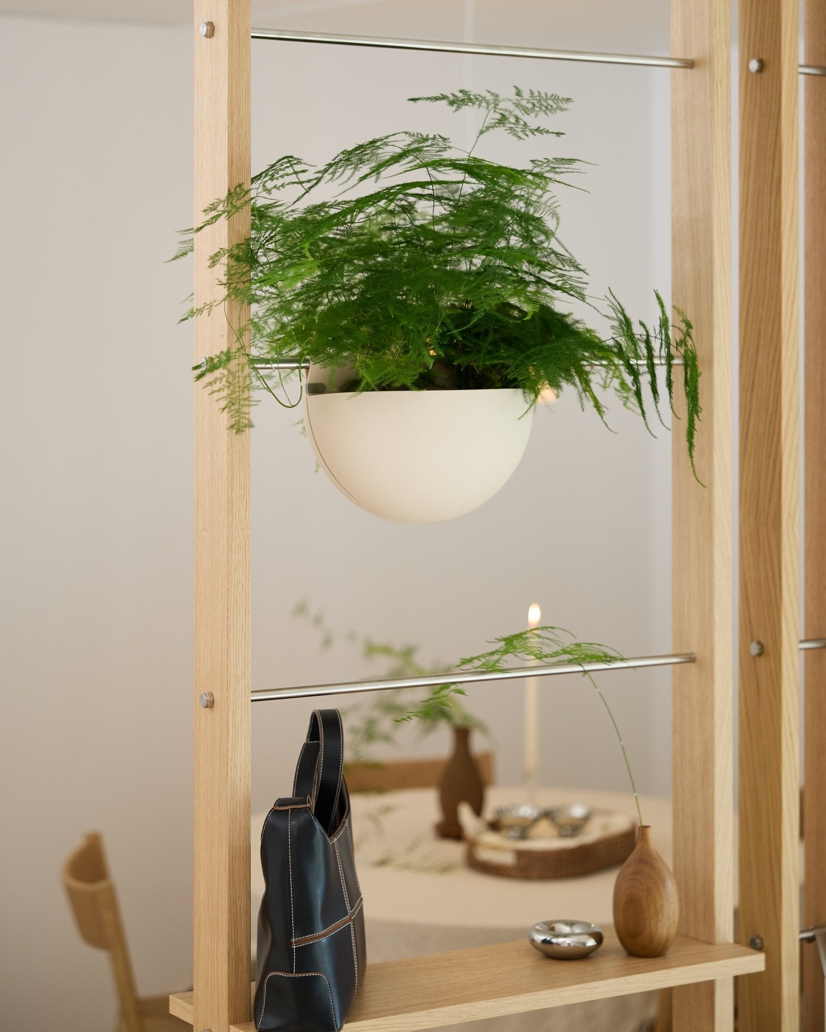 En grön ormbunke i en ljusbeige WoodUpp Gro Soft Pocket BM2-kruka står på en trähylla ovanför en svart väska, en vas och ett dukat matbord.