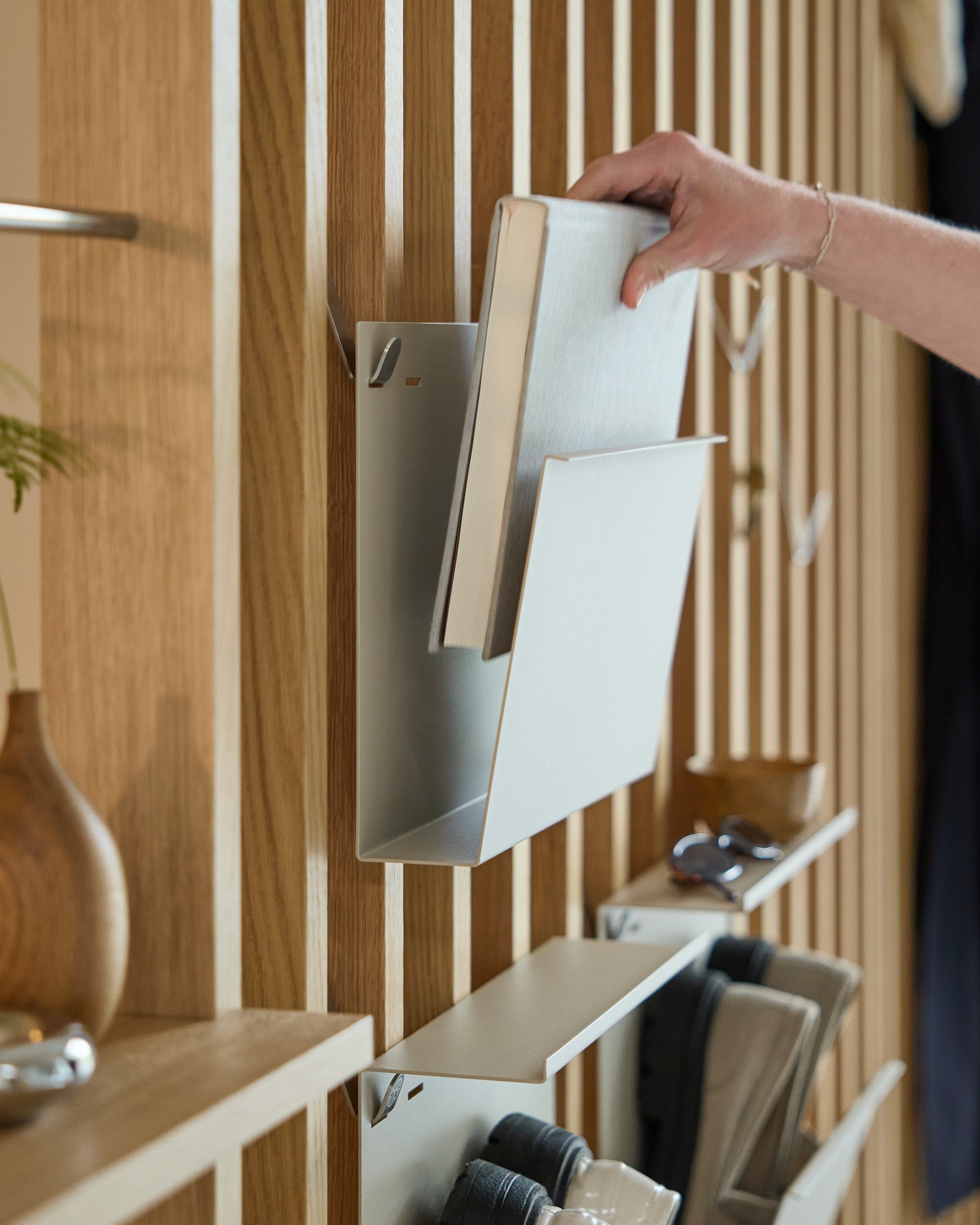 En hand placerar Nell | Magazine Storage / Beige från WoodUpp på en pulverlackerad steel hylla monterad på en nordisk trä-Akustikpanel, med andra föremål i närheten.