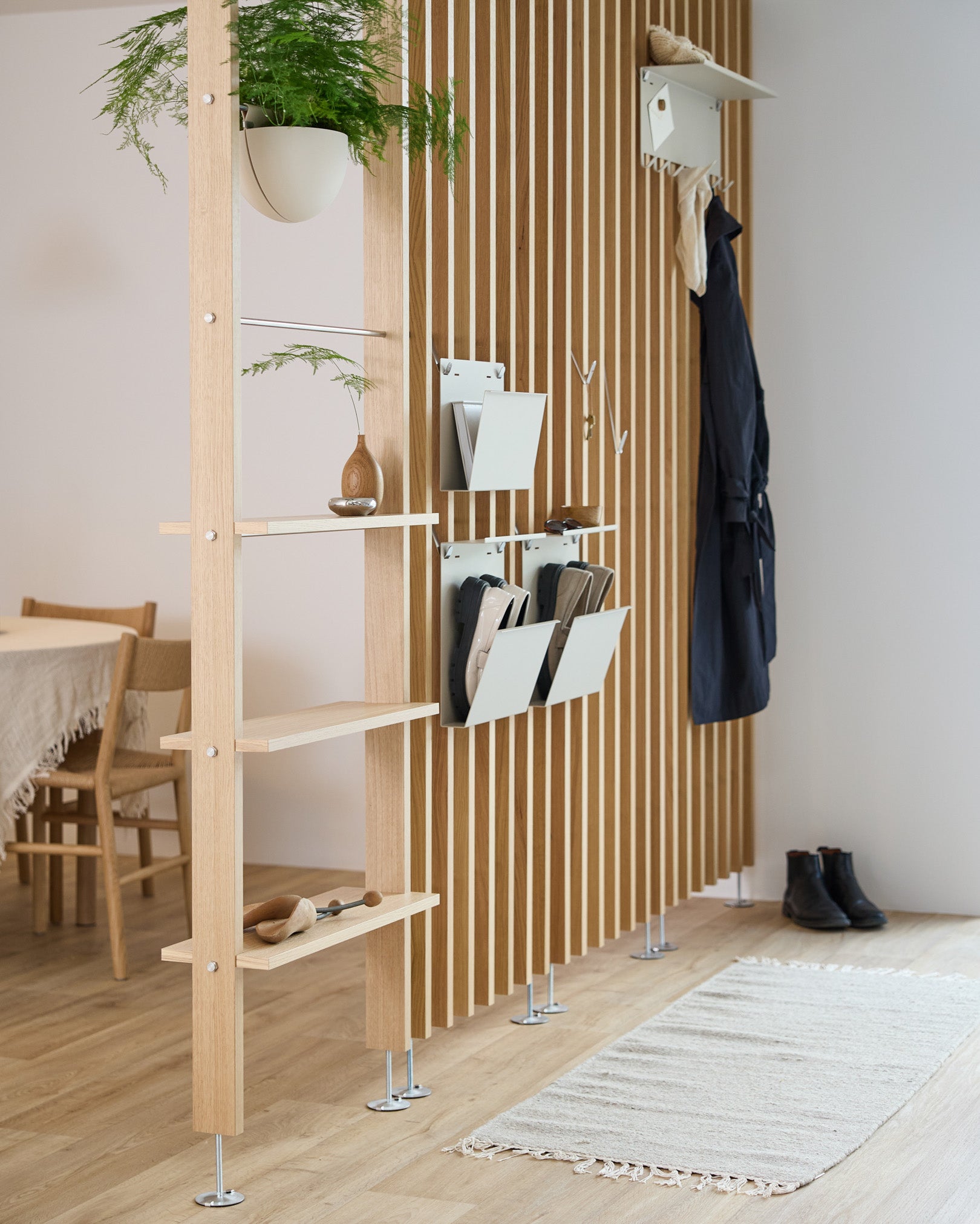 WoodUpp Nell Magazine Storage i beige integrerad i en rumsavdelare med steel hyllor, förvaringslådor och kappkrokar; matplats och matta syns bakom.