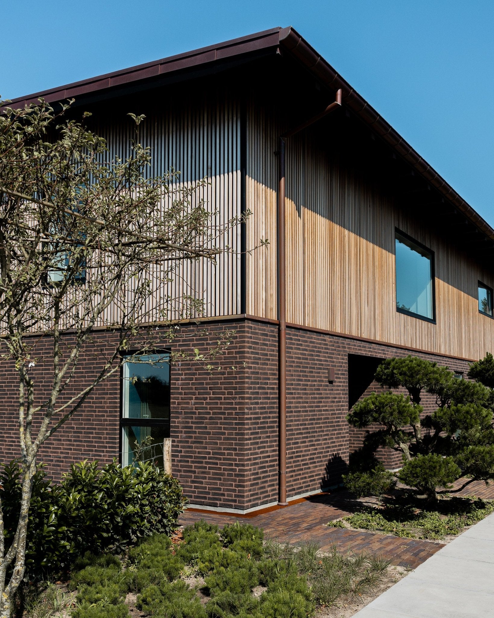 Two-story house with a brick lower level and vertical wood facade panels, surrounded by greenery under a clear blue sky.