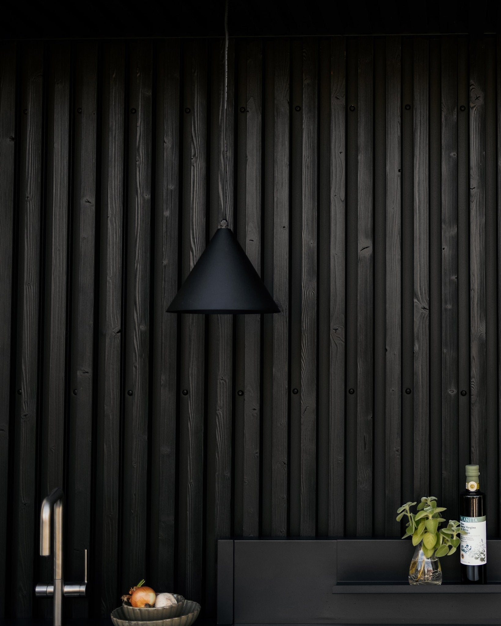 Modernt kök med matt black pendellampa, texturerad black Akupanel akustisk trävägg och bänkskiva med grönsaker, färsk basilika och olivolja.