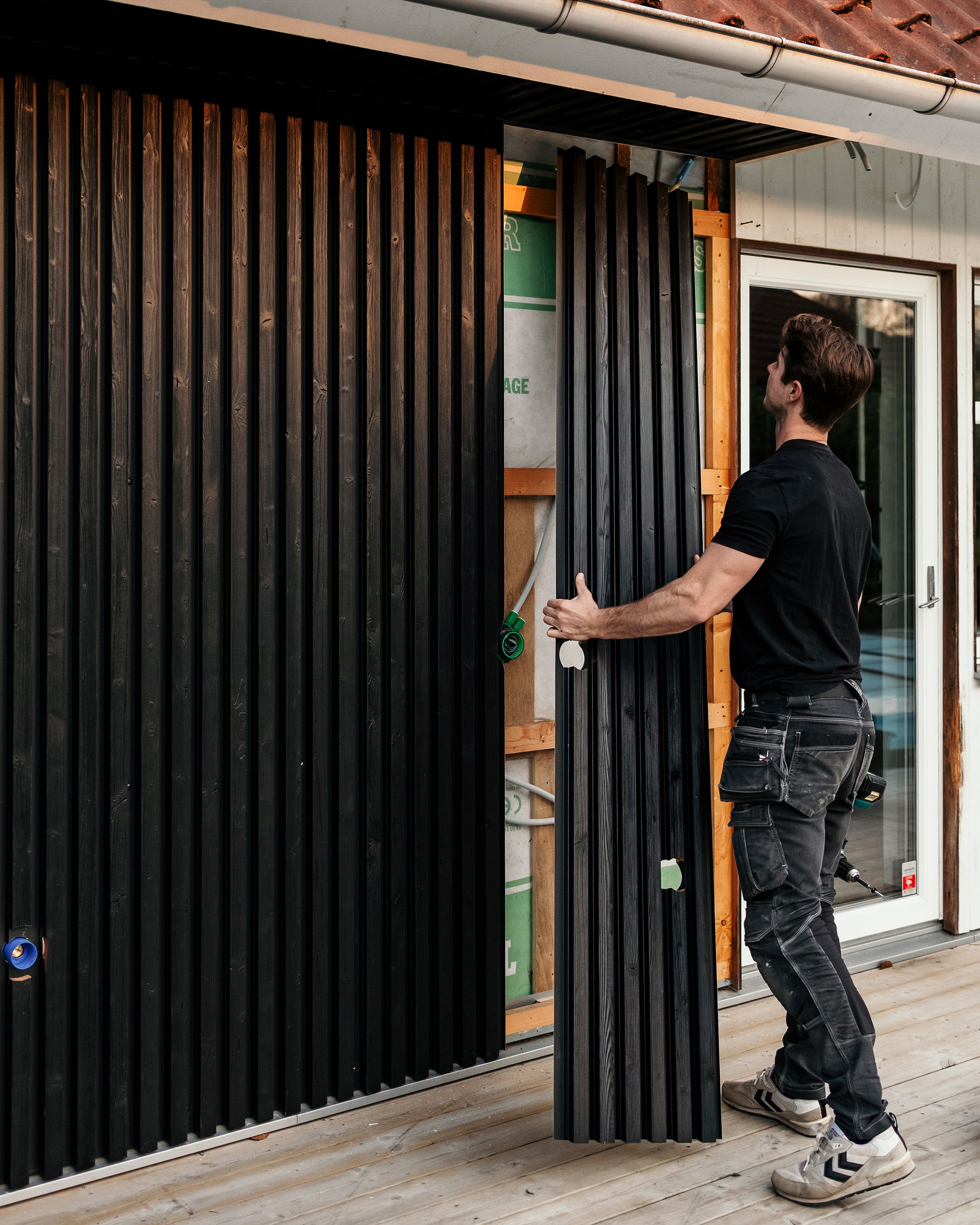 A man installs black vertical Aluwood facade panels on the exterior wall of a house under construction, working carefully on a wooden deck.