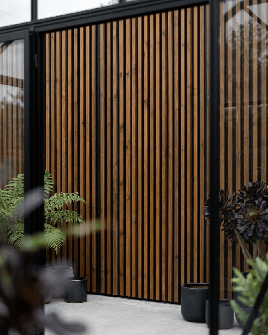WoodUpp Aluwood-fasadpanel för utomhusbruk i oljad brown, kombinerad med black krukor och gröna växter, som skapar en minimalistisk nordisk uteplats.