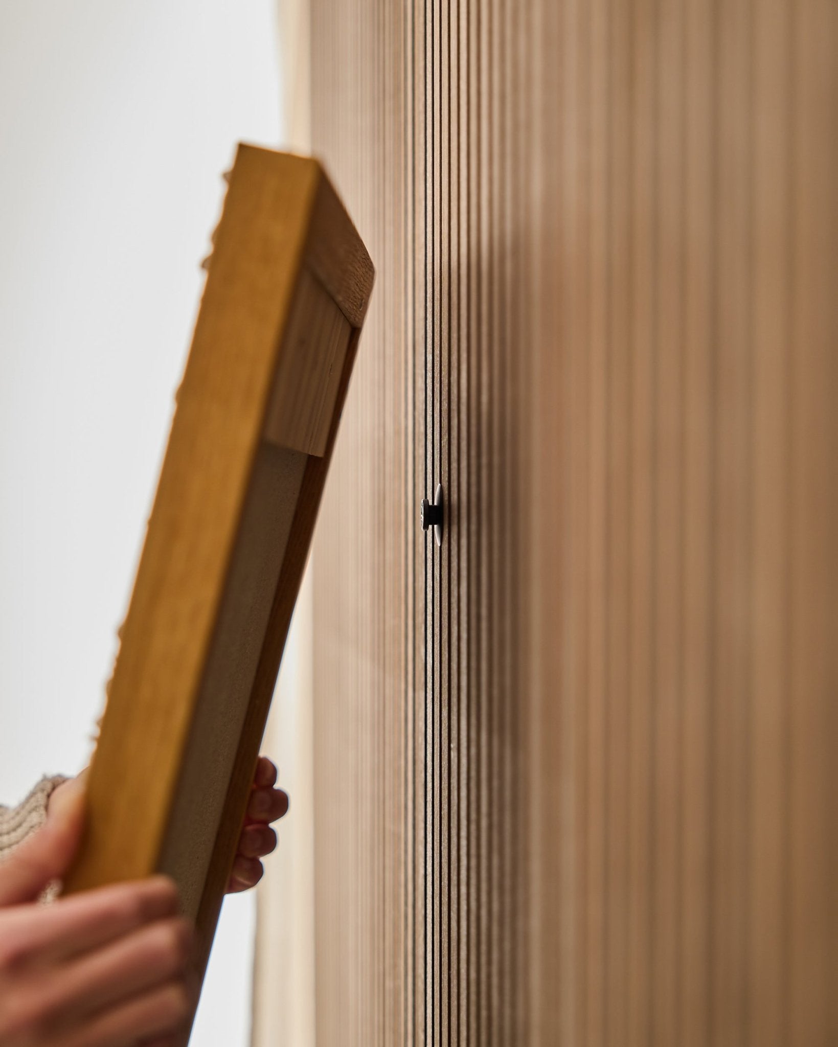 En person använder WoodUpp Dupps | Original-set för att montera en ram till en Akupanel akustisk träpanel, med fokus på att fästa en small black krok nära vertical ribbor.