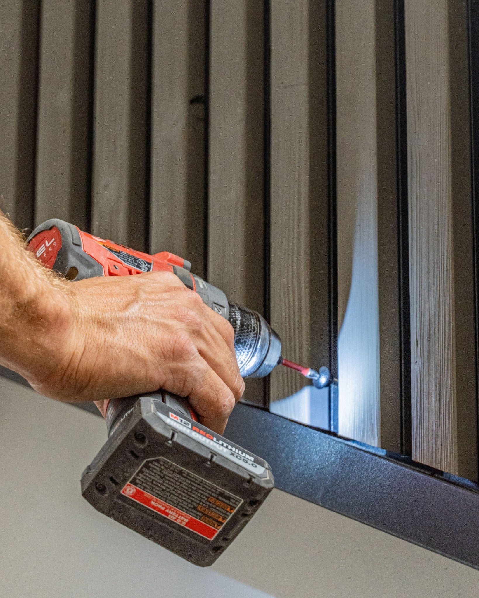 En person fäster en black WoodUpp Front Screw (4,8x35, Ø9,5-bricka) i en vertical Aluwood fasadpanel med en batteridriven borrmaskin.