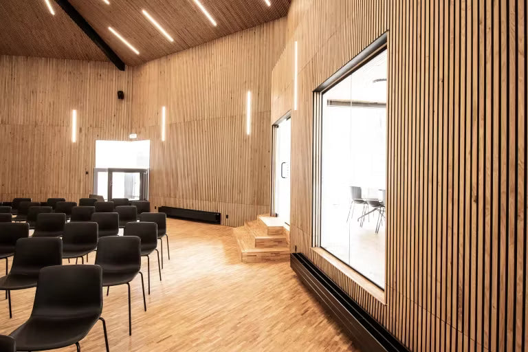 Rymligt konferensrum med Akupanel akustiska träpaneler på väggar och i tak, rader av svarta stolar, stora fönster och vertikala ljusarmaturer.