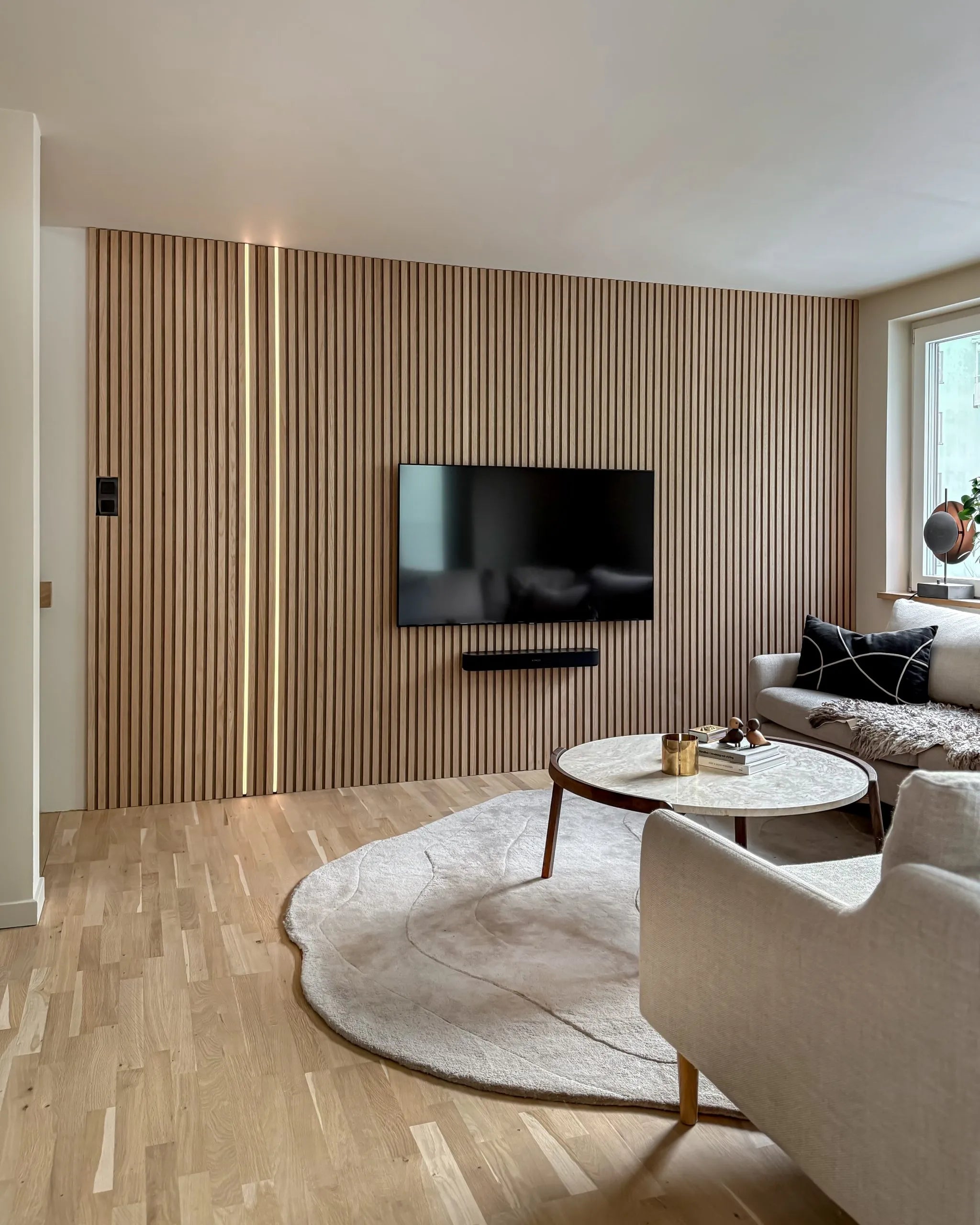 Modernt vardagsrum med en accentvägg av akustiska träribbor från Akupanel, monterad TV, runt soffbord, cremefärgad soffa, stor matta på ljust trägolv.