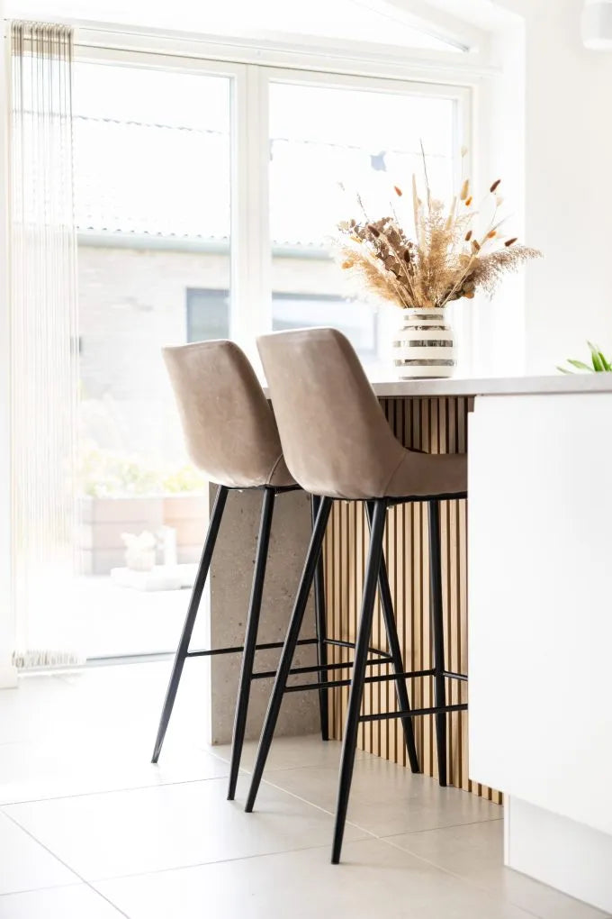 Beige barstolar vid en köksö med en vas med torkade blommor, stora fönster och nordisk modern stil med akustikpaneler på väggarna.