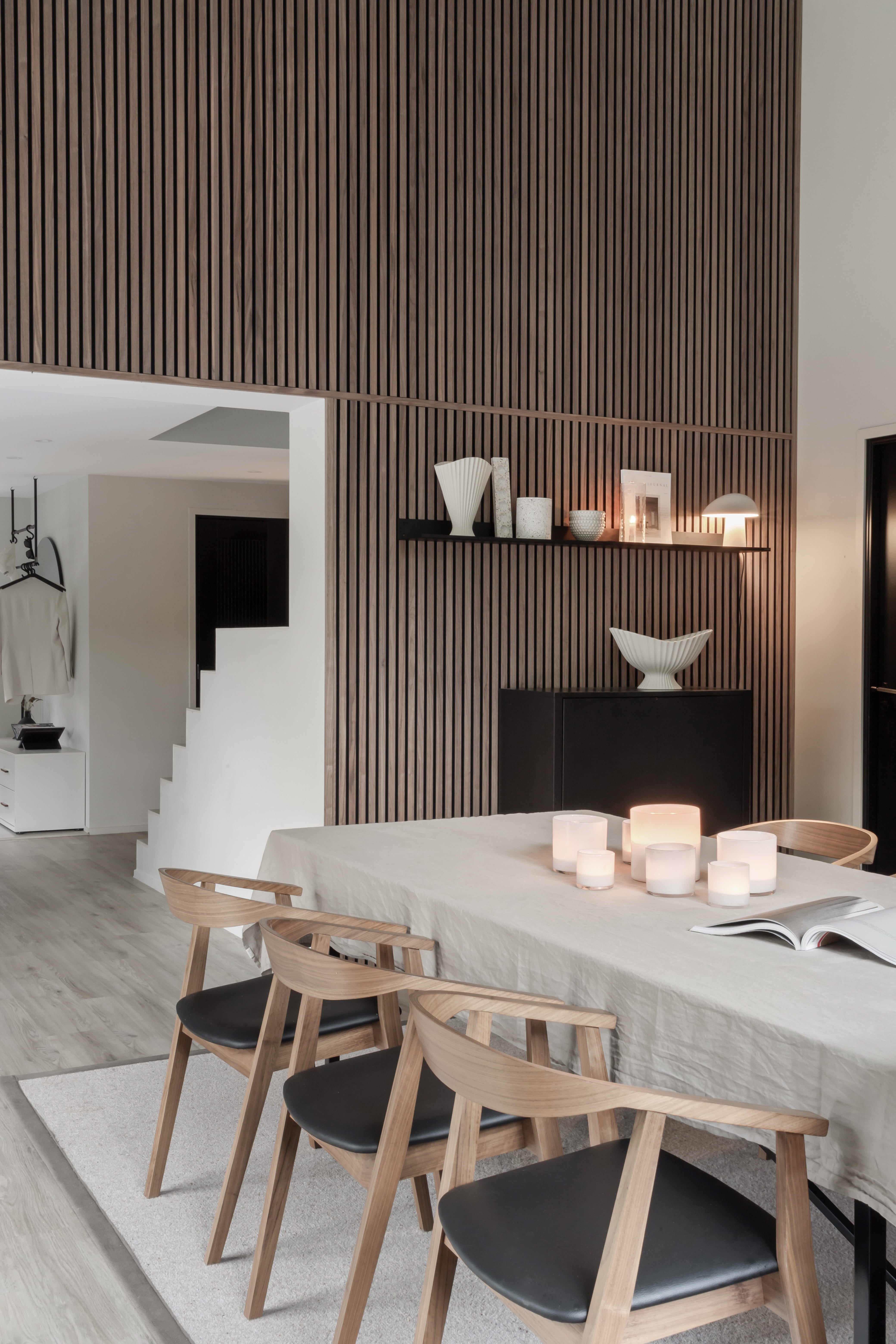 Modern matsal med bord i ljust trä, fyra stolar, levande ljus och minimalistisk inredning. Vertikal Akupanel akustisk träribbvägg och öppen trappa i utsikt.