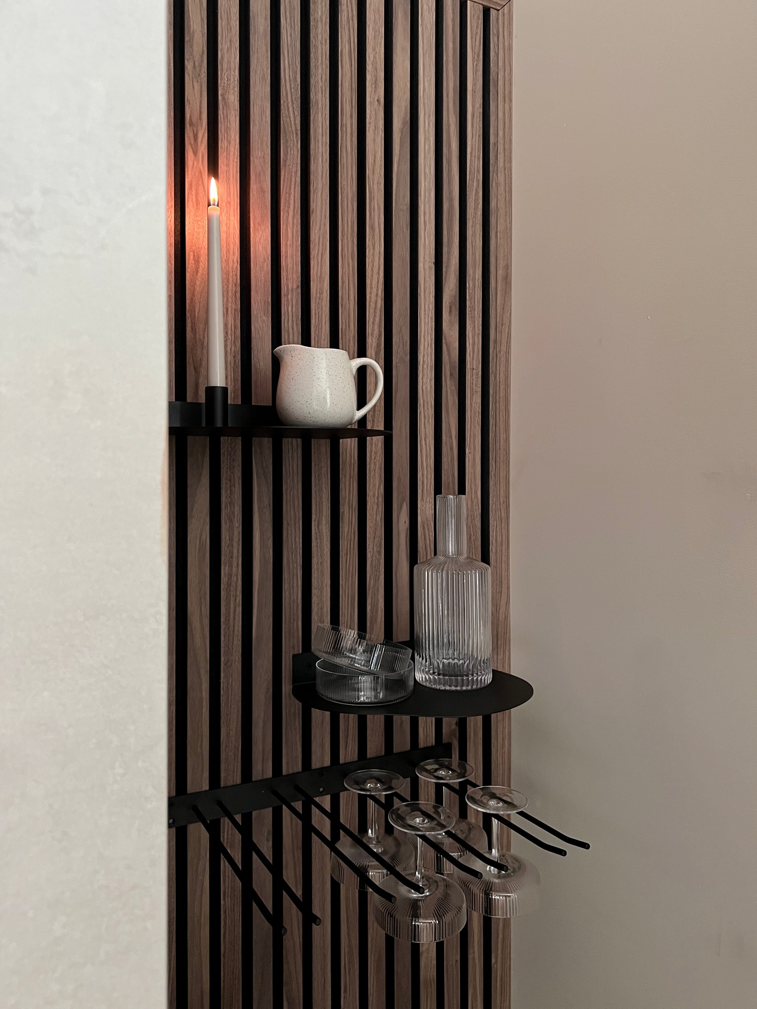 Modern nordisk akustisk slatwallpanel med svarta hyllor med ett tänt ljus, vit kanna, glasflaska och vinglas med stjälk hängande nedanför.