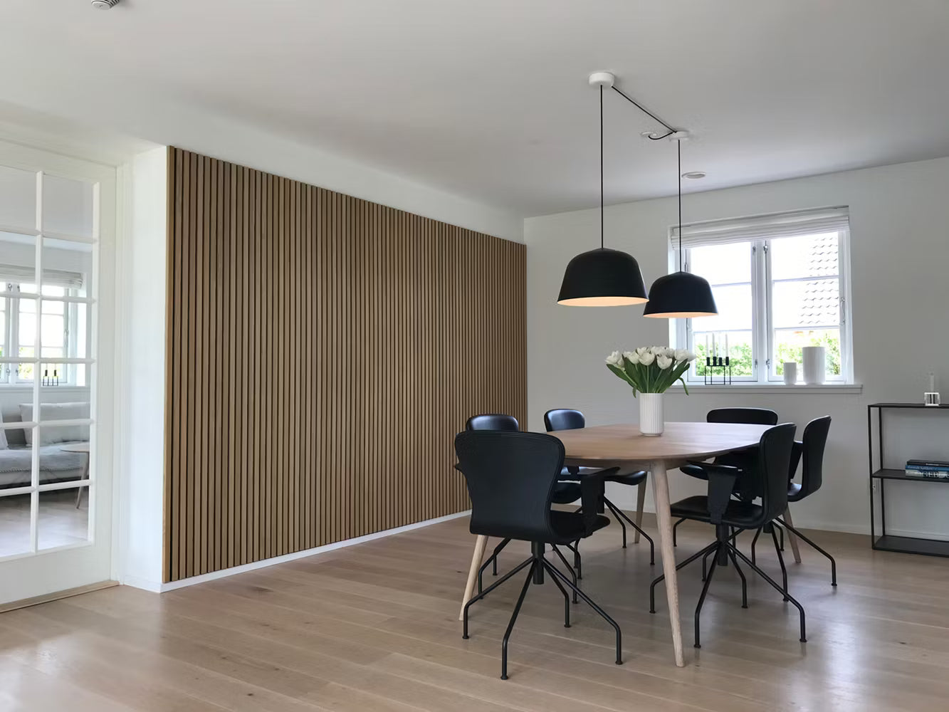 Modern matsal med trägolv, Akupanel akustiska lamellväggar, svarta stolar, träbord, pendellampor och en vit blommig mittpunkt.