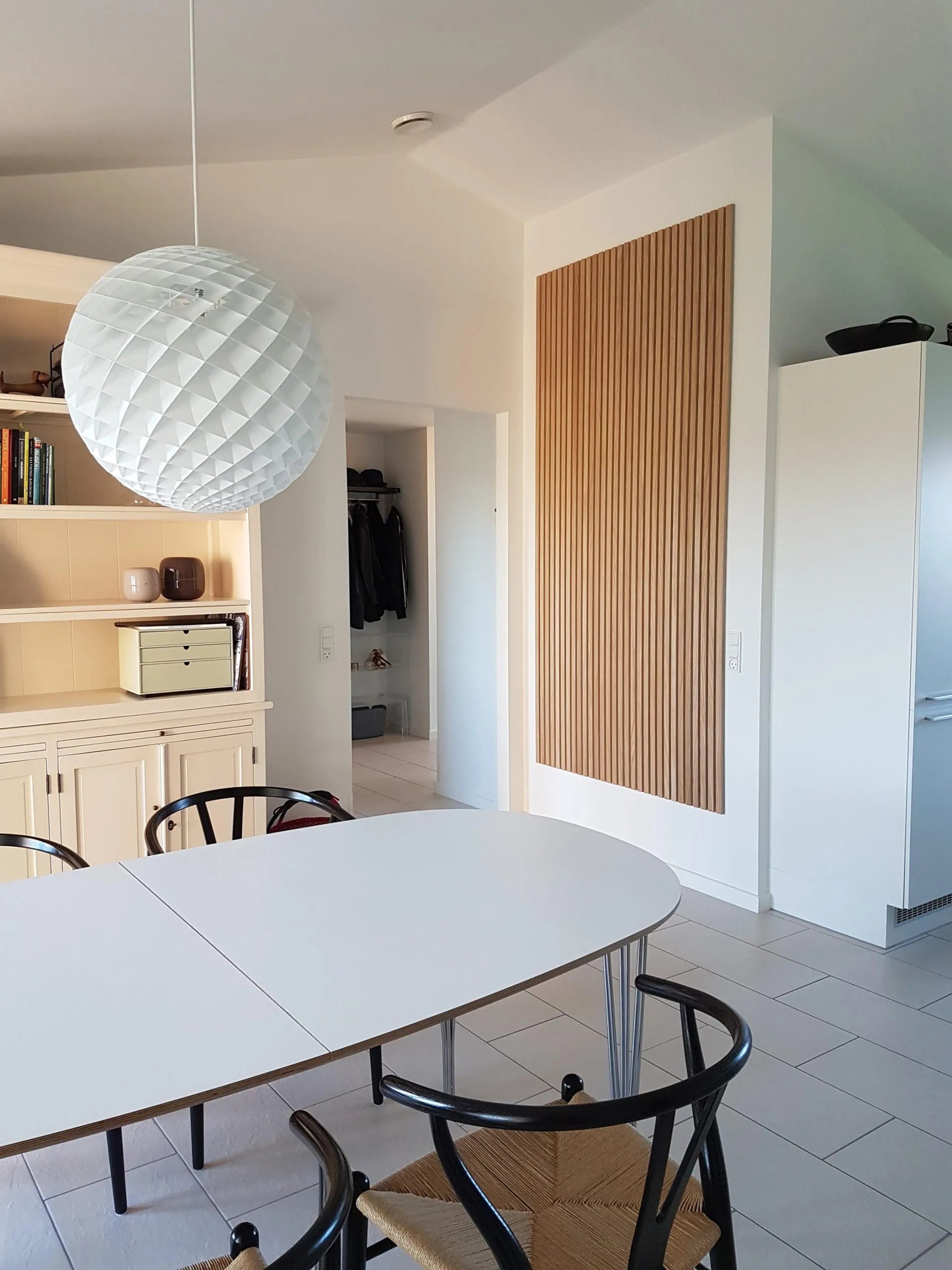 Modern matplats med ett vitt ovalt bord, svarta stolar, stor texturerad taklampa och snygga Akupanel-skåp i ljust trä.