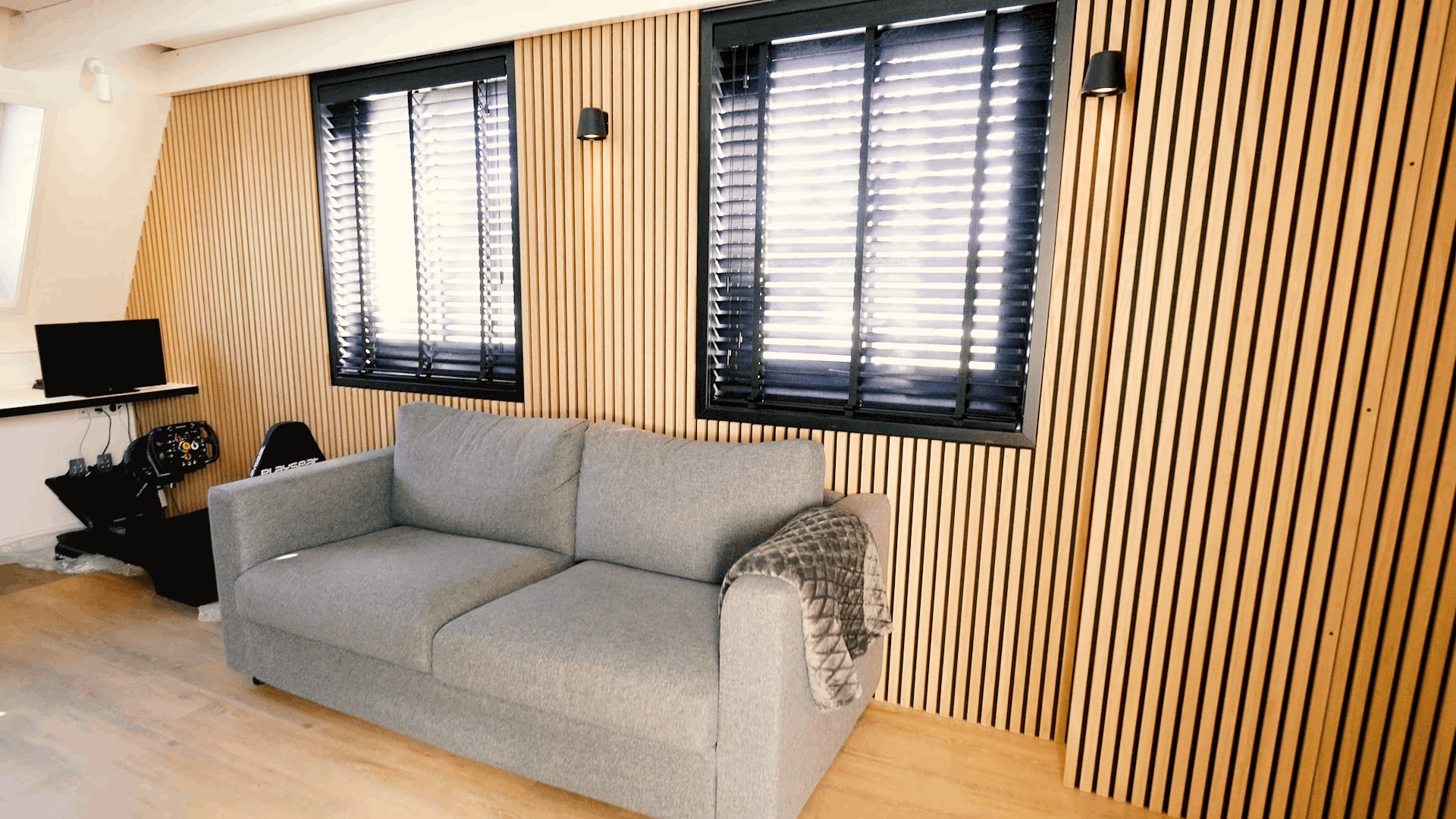 Grå soffa med mönstrad filt placerad framför en nordisk Akupanel akustisk träribbvägg, flankerad av två fönster med svarta persienner och en kompakt skrivbordsuppställning.