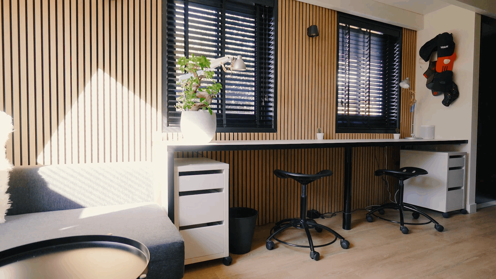 Modernt kontor med nordiska akustiska träpaneler på väggarna, ergonomiska arbetsstationer, svarta stolar, en krukväxt och solljus genom svarta persienner.