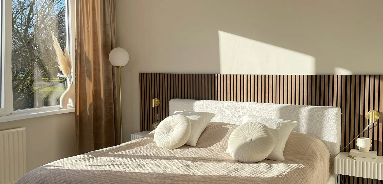 Solbelyst modernt sovrum med en beige quiltad säng, runda kuddar, Akupanel sänggavel med träribbor och minimal inredning intill ett stort fönster med skira gardiner.