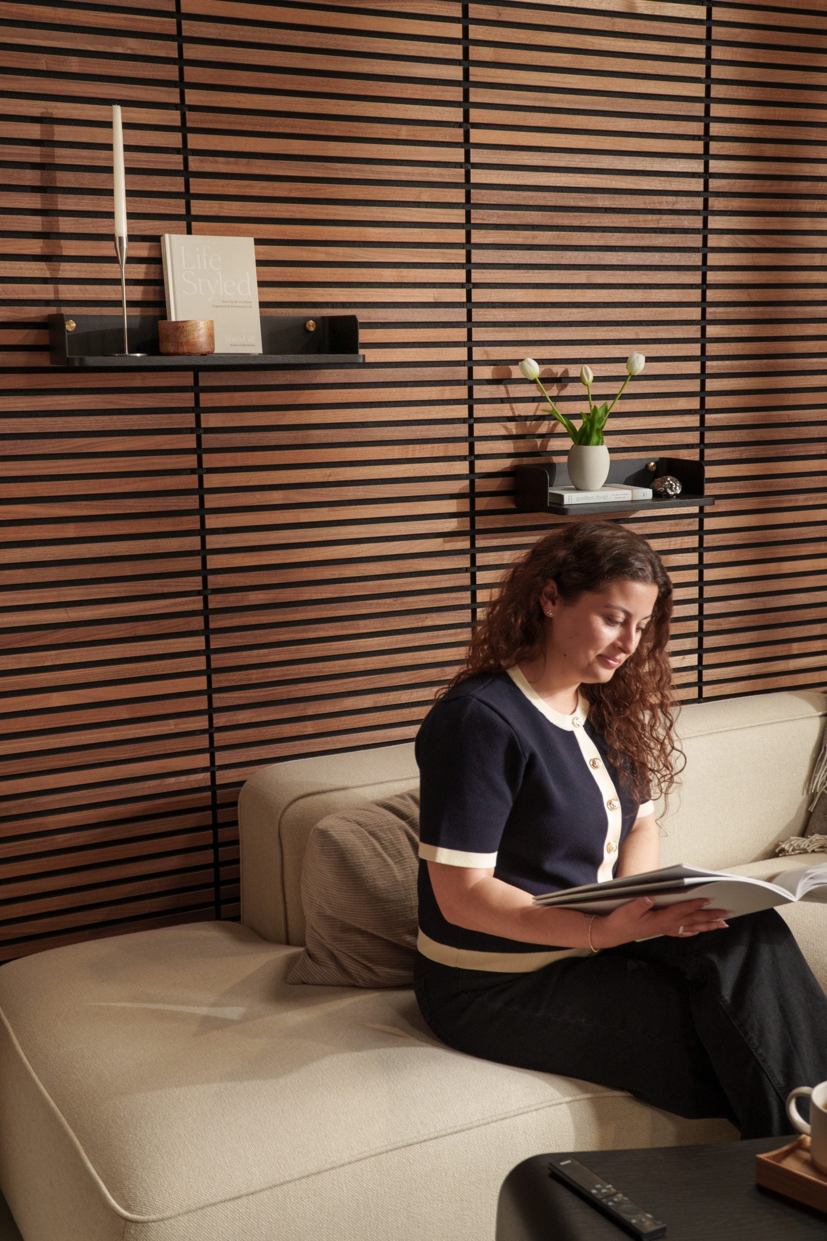 A curly-haired woman relaxes on a beige sofa reading, surrounded by Nordic Akupanel acoustic wood slatwall panels, shelves, and a small plant in the background.