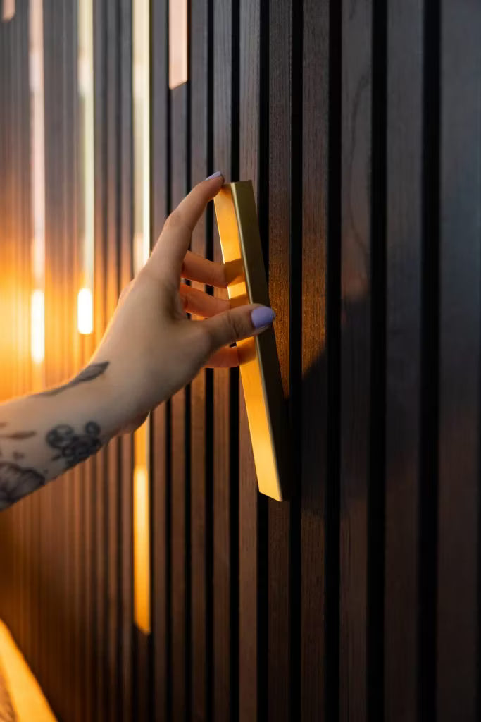 En tatuerad hand med ljuslila naglar greppar ett guldhandtag på en elegant, mörk Akupanel akustisk träpanelvägg, framhävd av varm belysning.