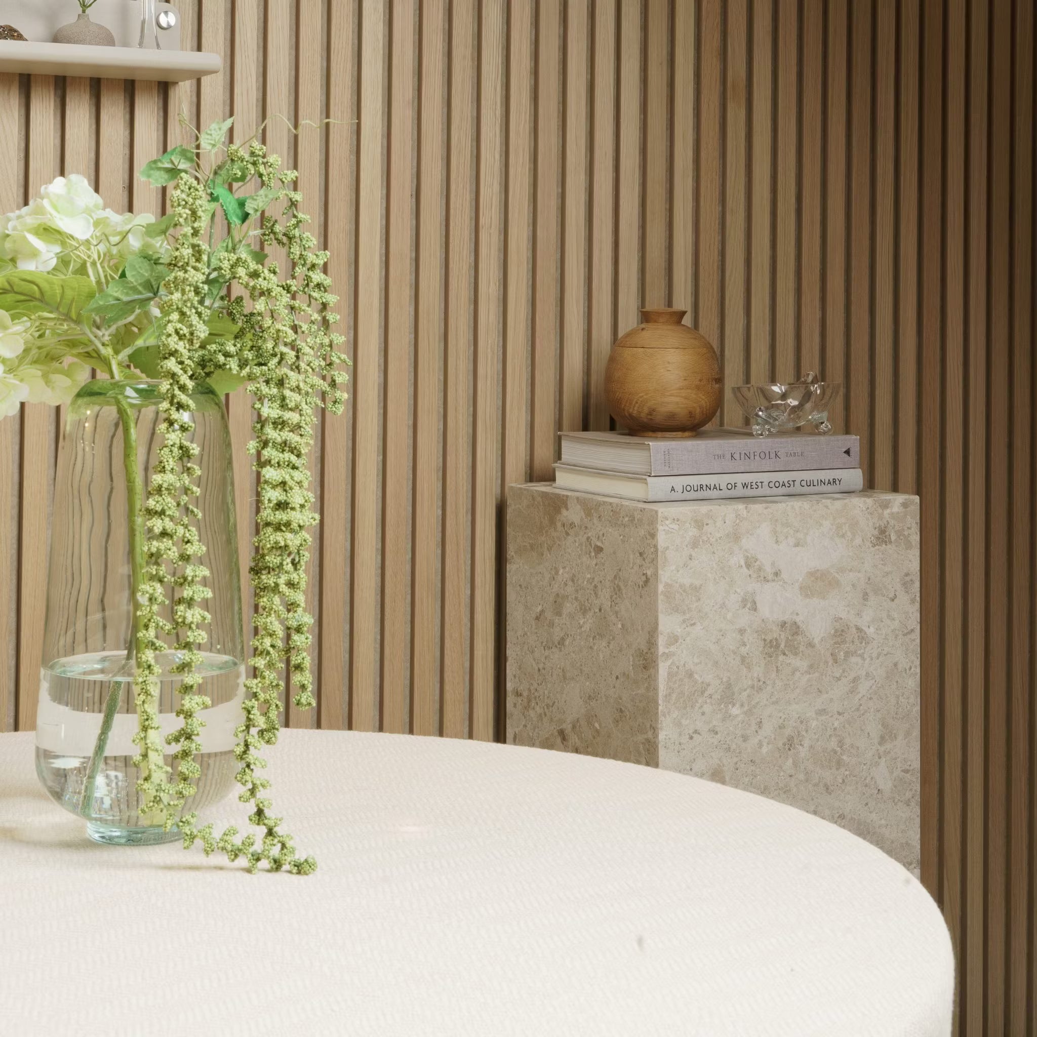 En vas med blommor står på ett runt bord, böcker och keramik vilar på en marmorpiedestal mot en vägg av Akupanel akustisk träpanel.
