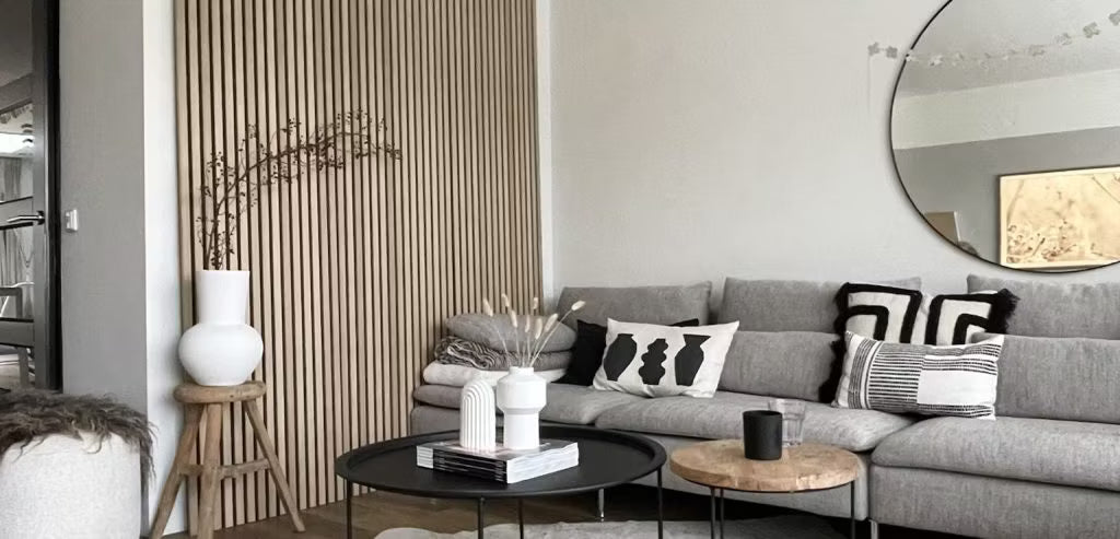Modernt vardagsrum med en grå soffa, abstrakta kuddar, svarta soffbord, dekorativa vaser och en Akupanel akustisk lamellvägg i trä med en rund spegel.